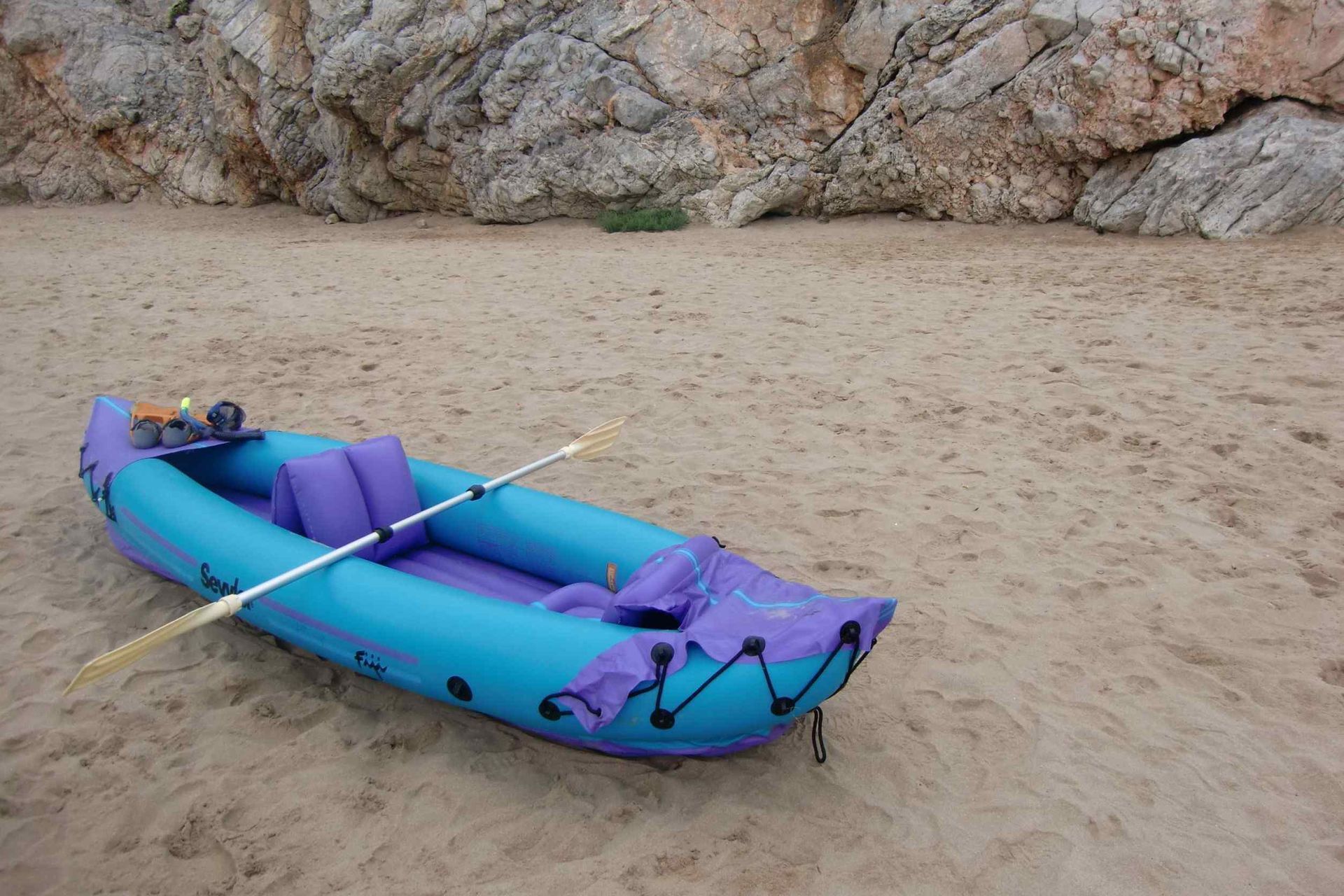 Un kayak inflable azul y morado con remos en una playa de arena.