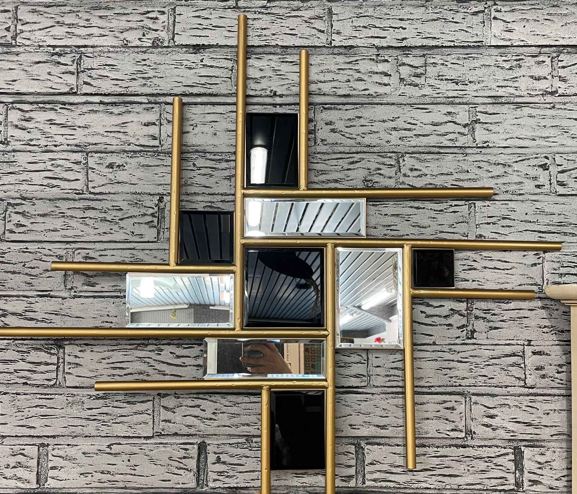 Miroir mural abstrait avec cadre doré et plusieurs sections en miroir, contre un mur de briques.