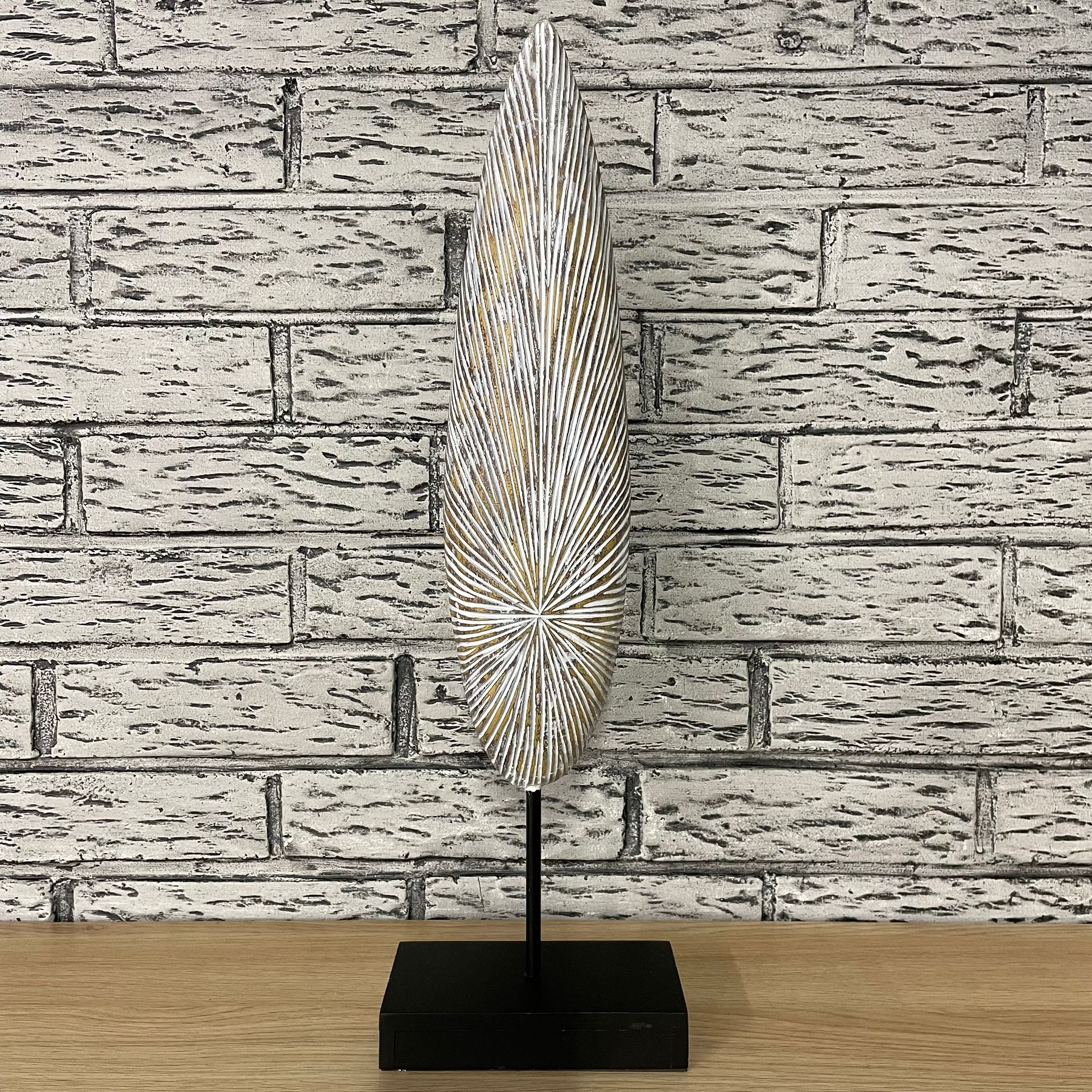 Sculpture abstraite en bois blanc et or sur un support noir, posée contre un mur de briques.