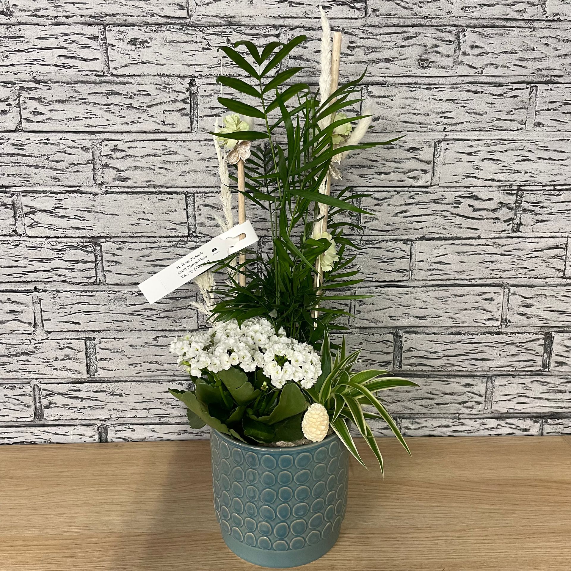 Composition florale dans un pot bleu avec des fleurs blanches, des plantes vertes, contre un mur de briques blanches.