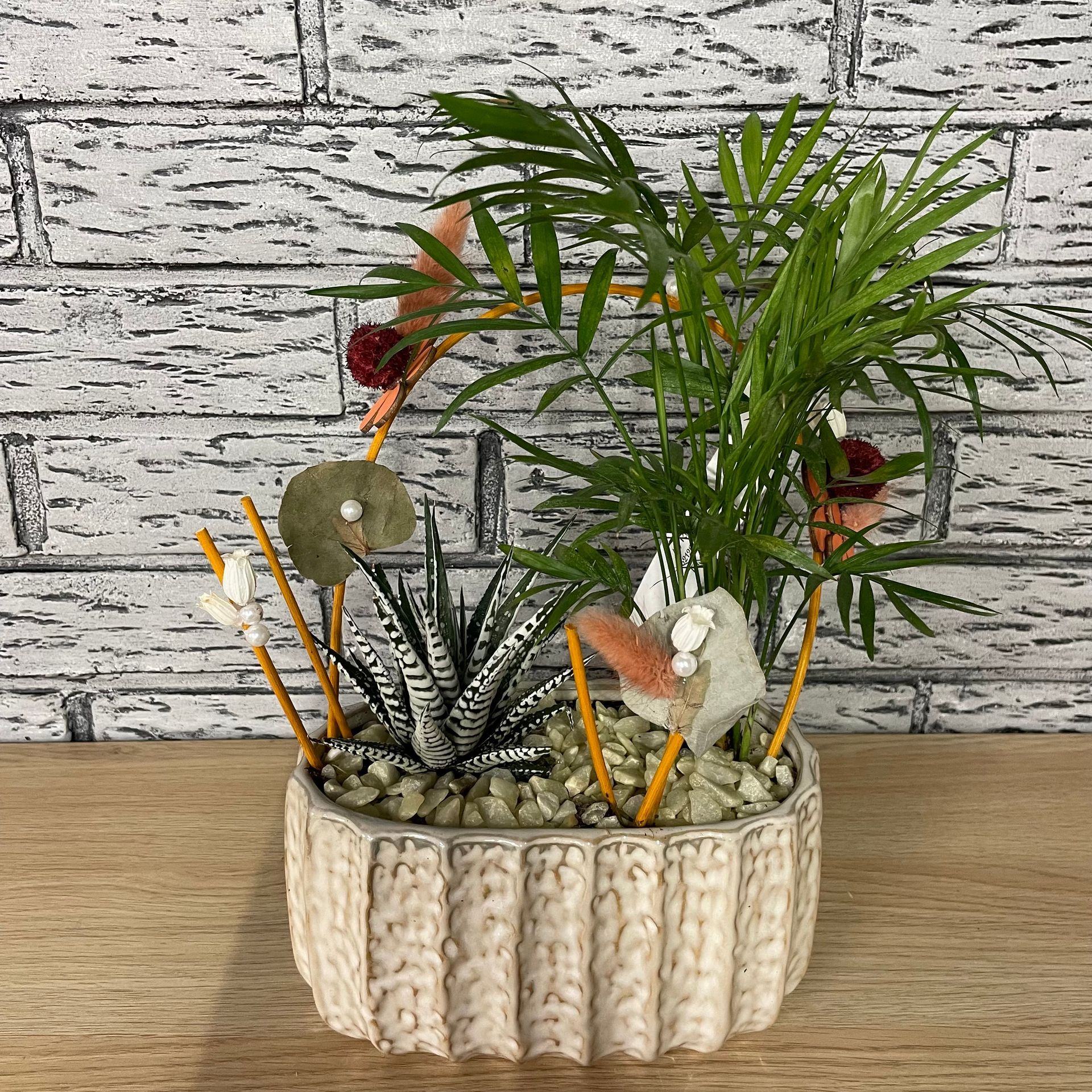 Plantes succulentes et palmiers disposés dans un pot texturé de couleur crème avec des éléments décoratifs contre un mur de briques.