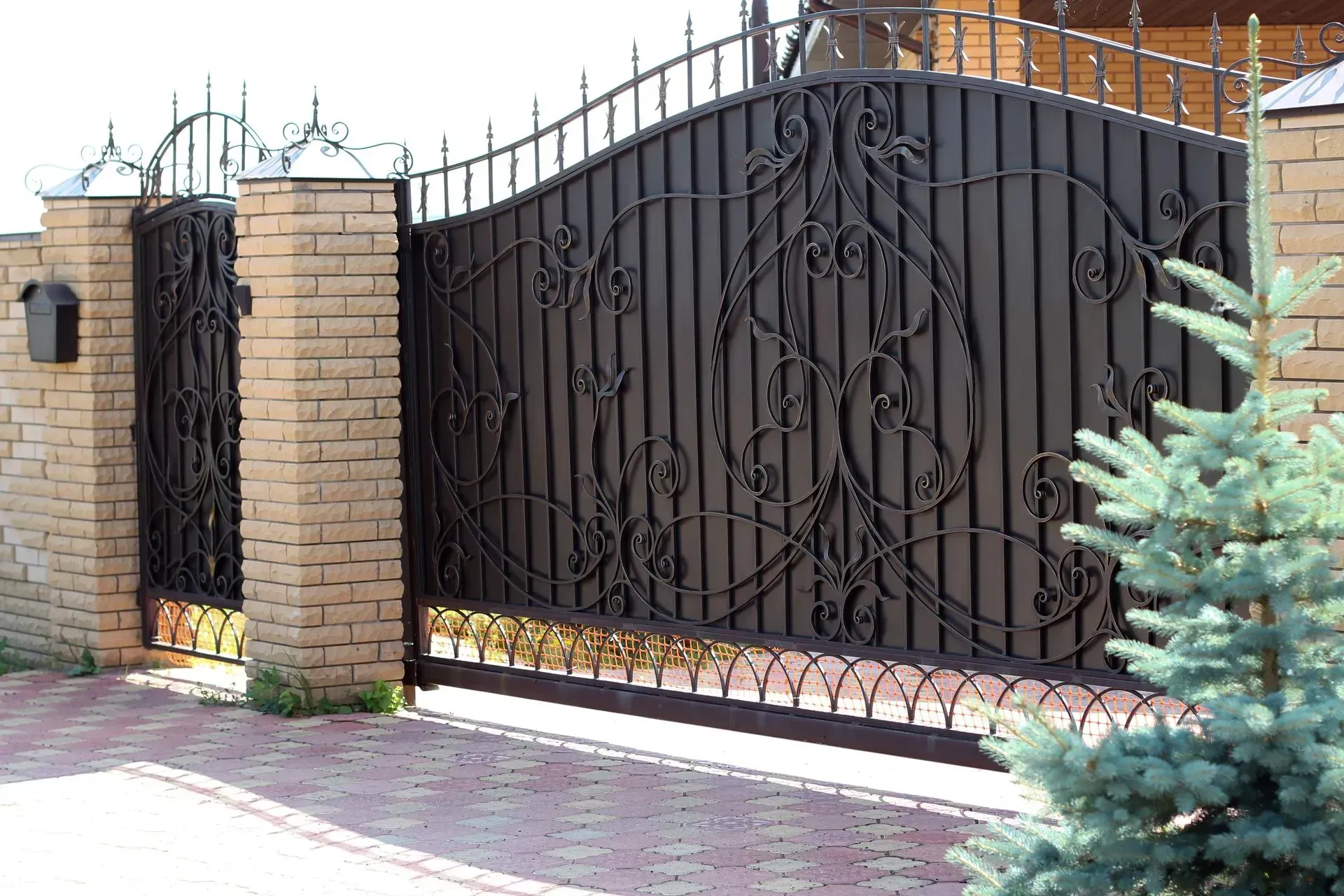 Puerta de hierro forjado negra con enredaderas decorativas entre pilares de ladrillo; frente de una casa.