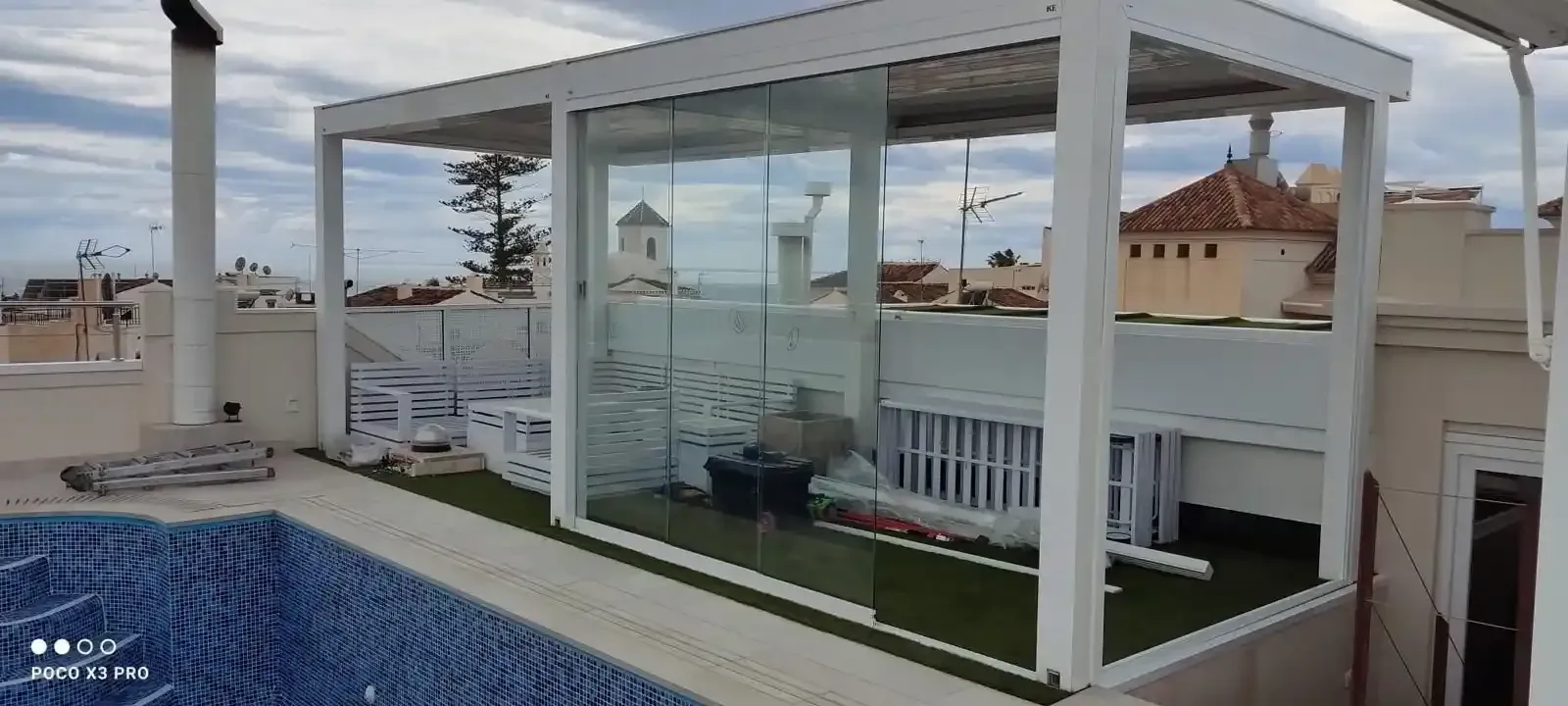 Patio en la azotea con piscina y zona de estar acristalada. Estructuras blancas, césped artificial y vistas a la ciudad.