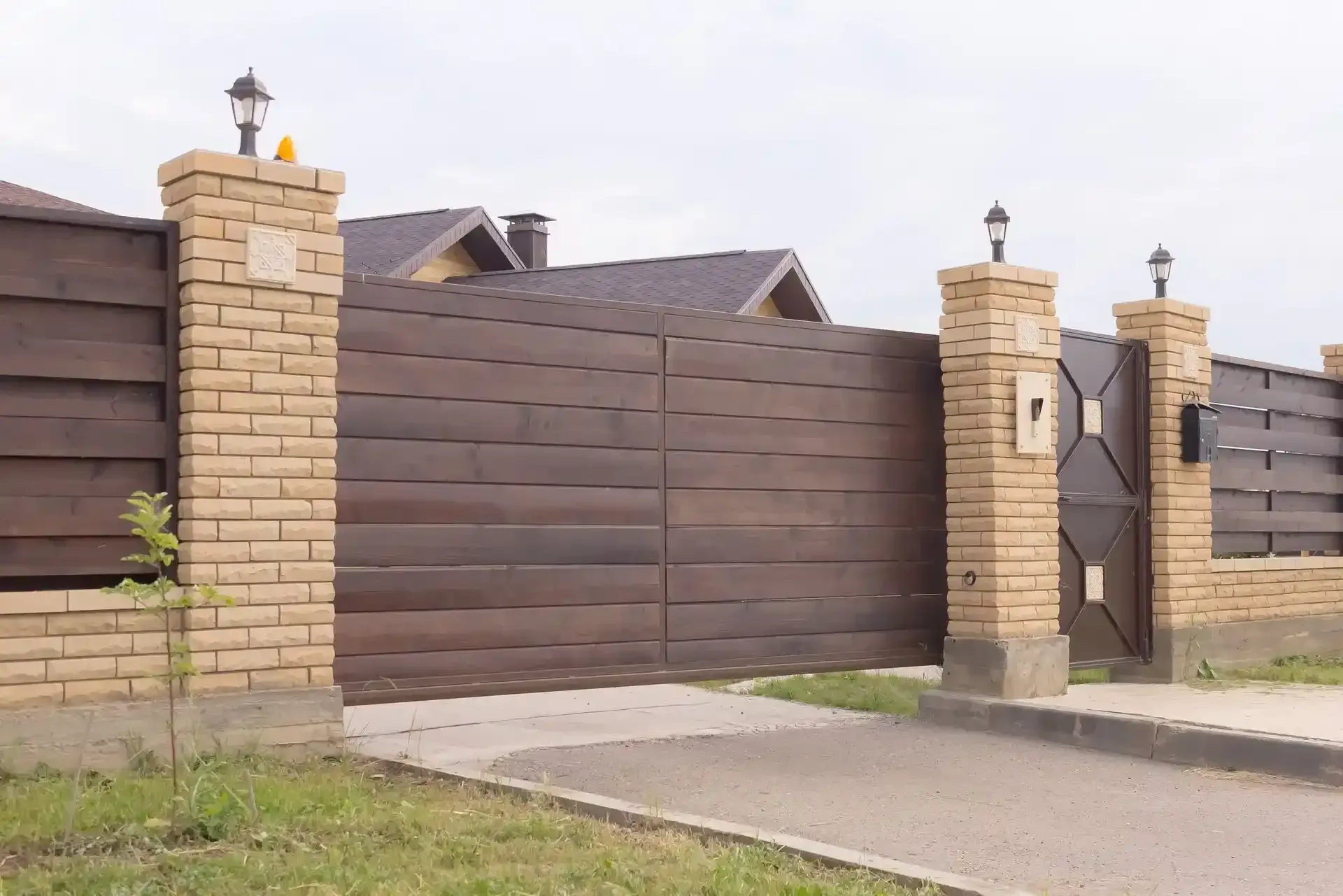 Puerta de madera y valla con pilares de ladrillo, que conduce a una casa, sobre un camino de entrada pavimentado.