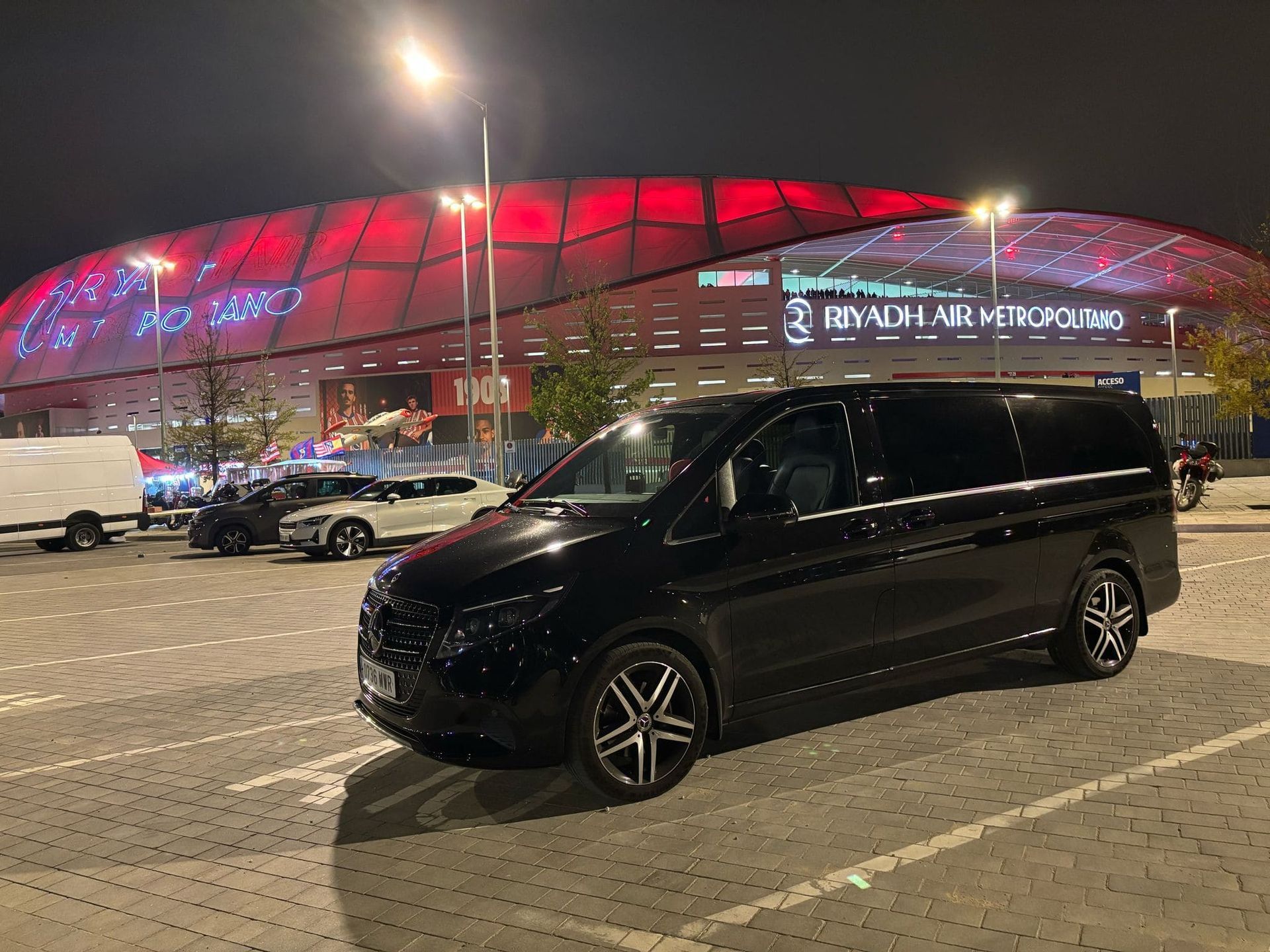 Camioneta negra estacionada frente a un estadio con el techo iluminado en rojo por la noche.