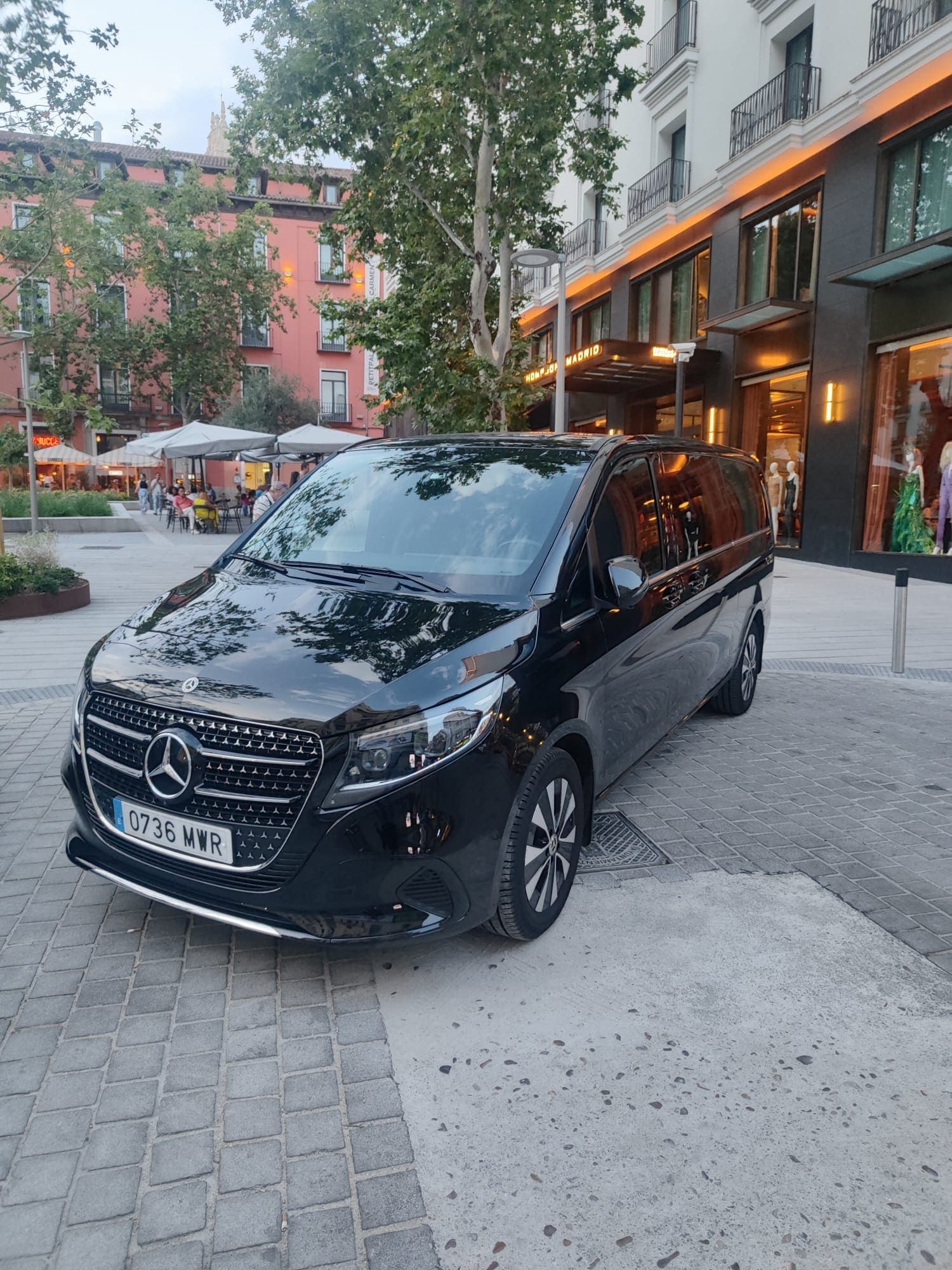 Minivan Mercedes negra estacionada en una calle adoquinada frente a un edificio con tiendas.