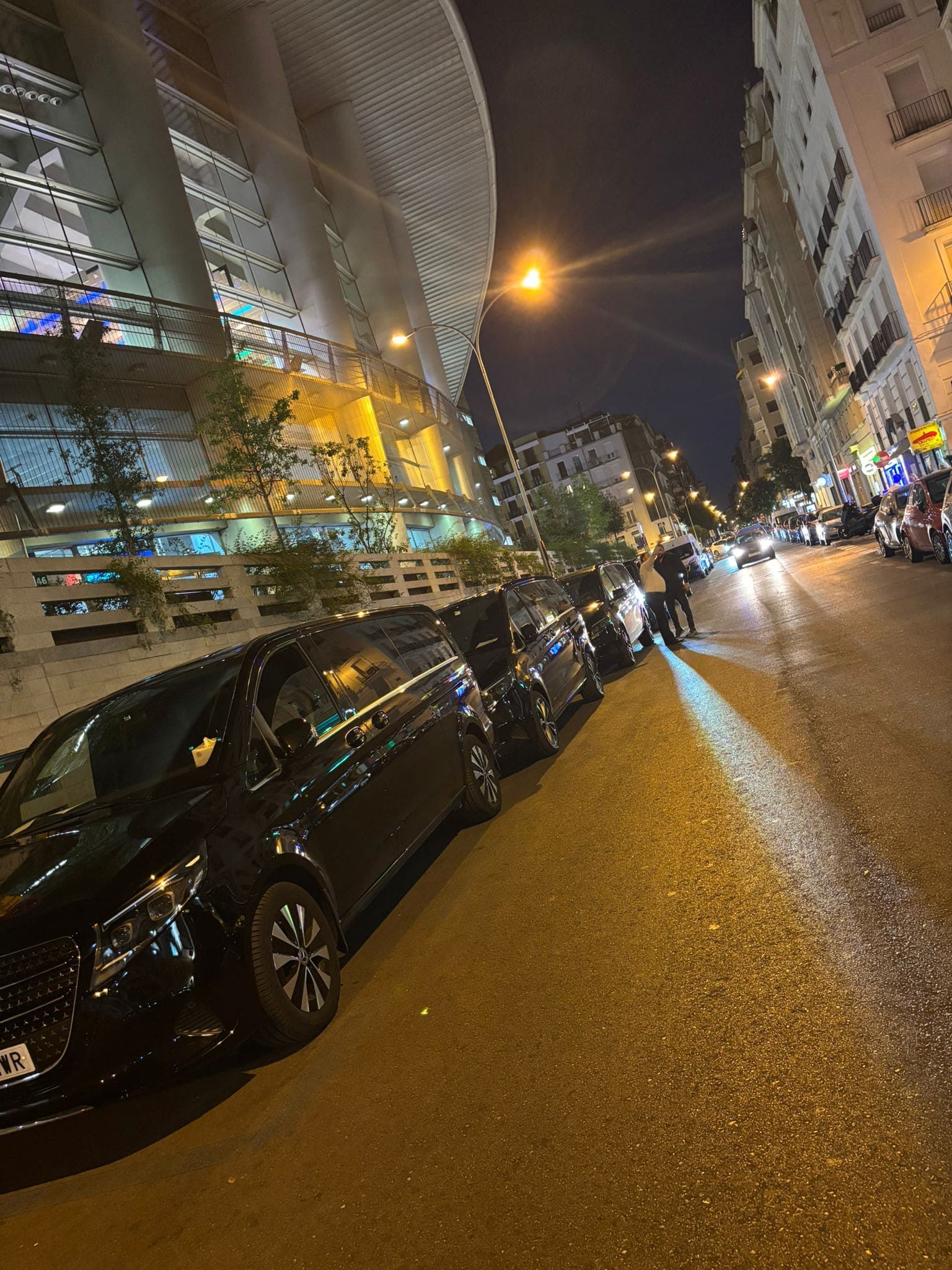 Calle de noche con coches aparcados, edificios y una estructura parecida a un estadio.