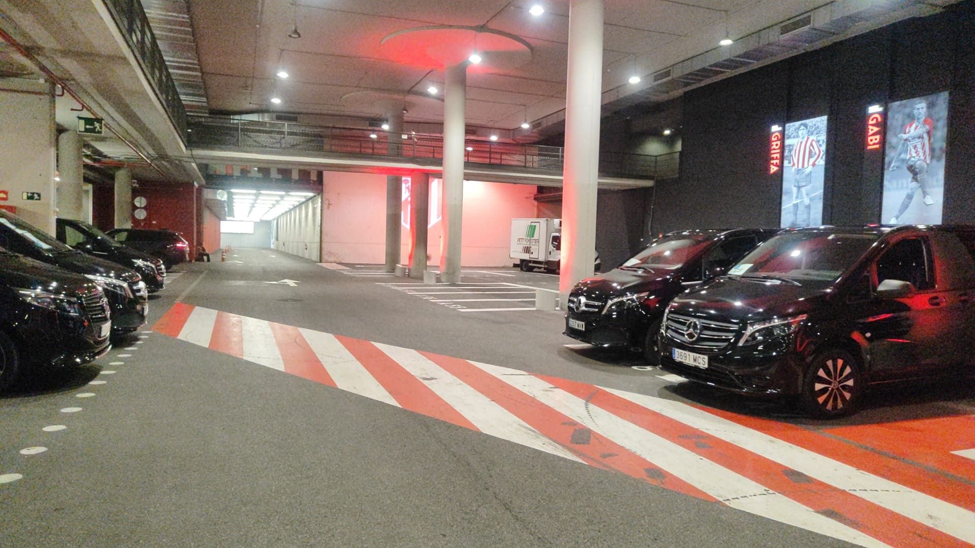 Aparcamiento subterráneo con varias furgonetas negras estacionadas en carriles con franjas rojas y blancas. Iluminación roja en el techo.