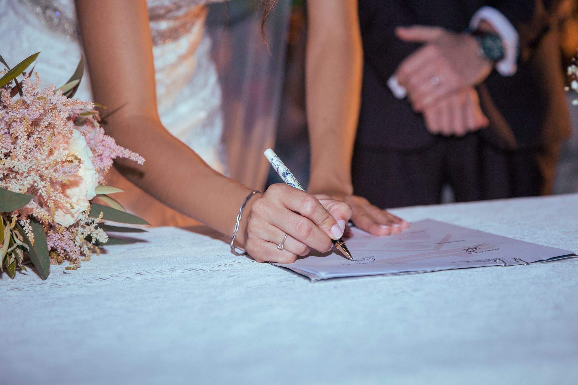 Lors d'une cérémonie de mariage, la mariée signe un document à la main, stylo à la main. Gros plan sur ses mains et le papier, le bras du marié étant visible.