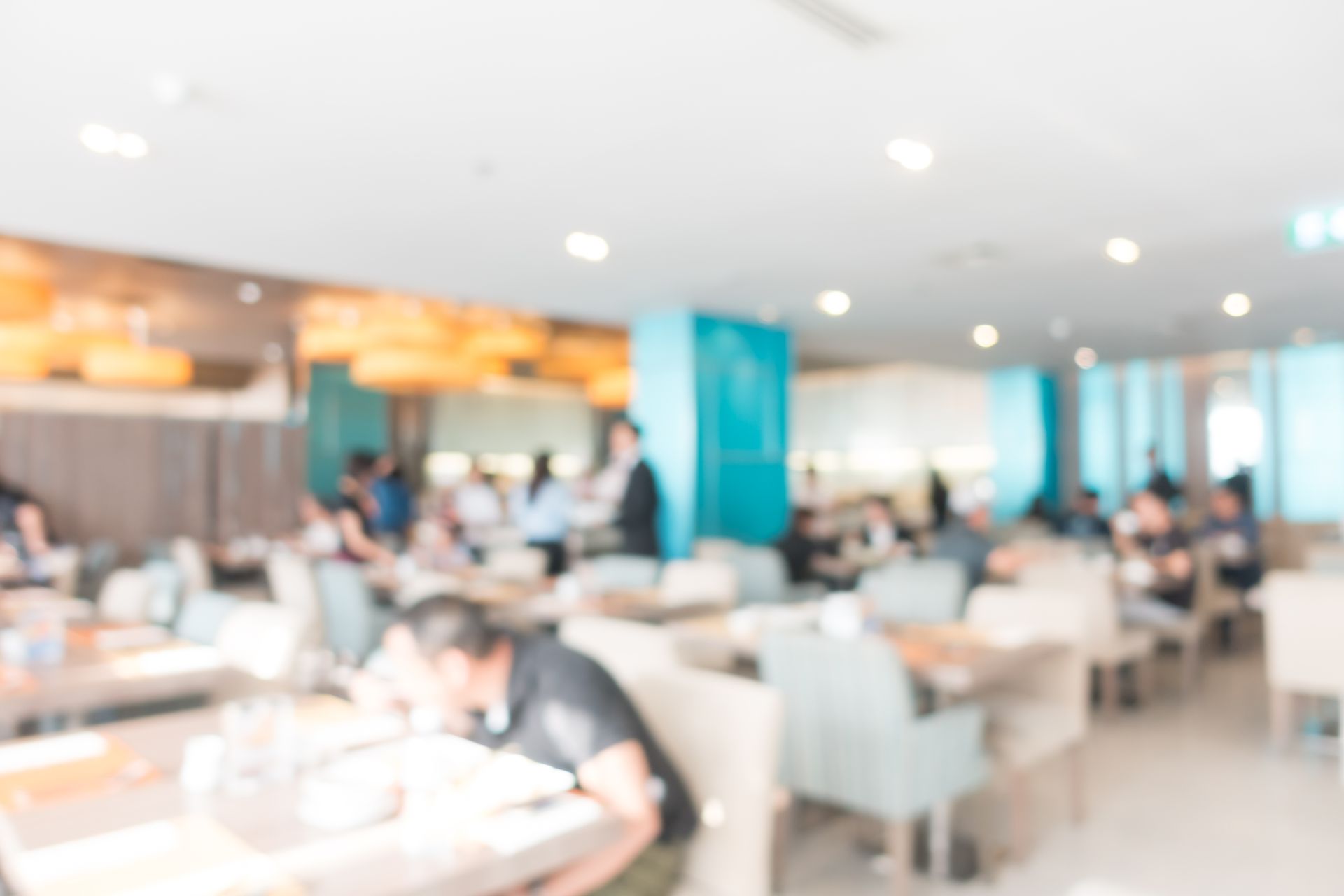 Vue floue d'une salle à manger lumineuse avec des personnes assises à des tables, un serveur et des touches turquoise.