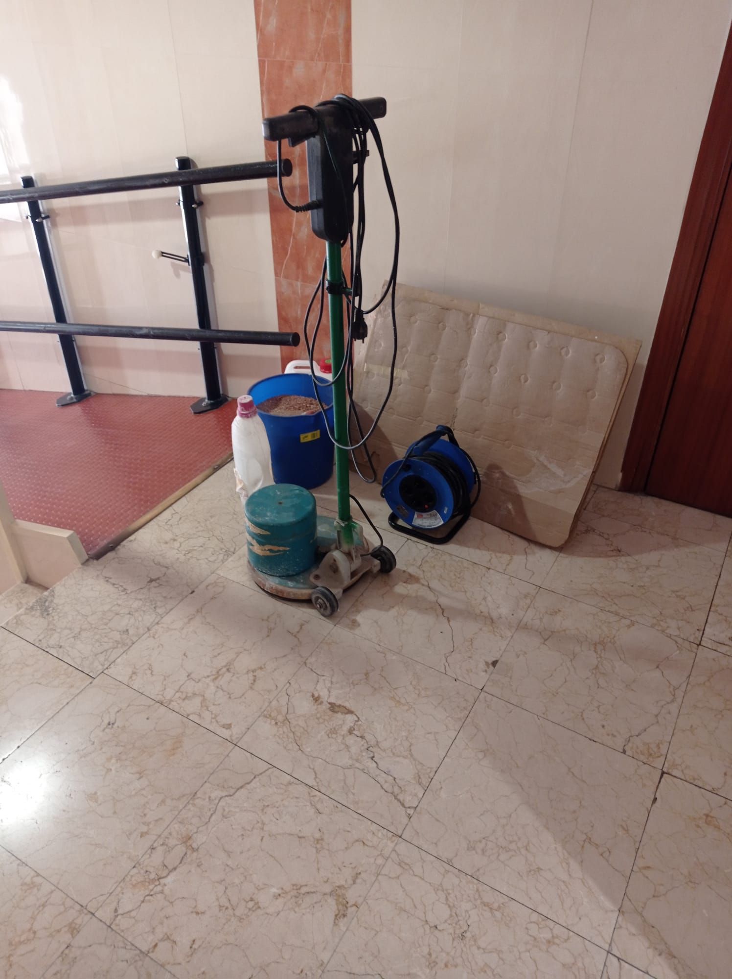 Una pulidora de suelos, un cubo azul y un colchón enrollado reposan sobre un suelo de baldosas cerca de una barandilla negra.
