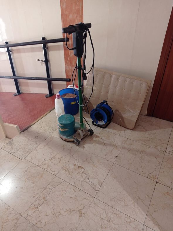 Una pulidora de suelos, un cubo azul y un colchón enrollado reposan sobre un suelo de baldosas cerca de una barandilla negra.