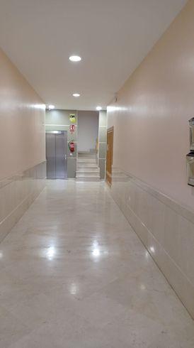 Un pasillo luminoso y vacío, con suelos y paredes de mármol, que conduce a un ascensor y a una corta escalera al final.