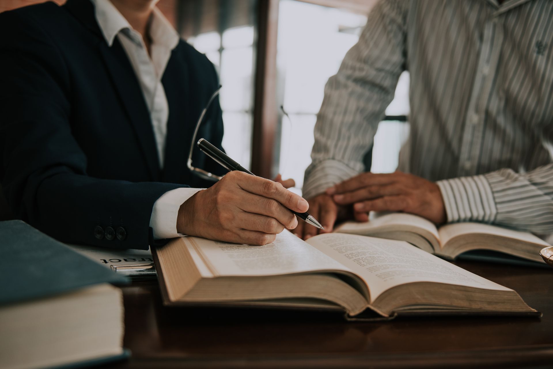 Une personne en costume écrit dans un grand livre ouvert, une autre personne se tient à proximité, des livres sont posés sur un bureau.