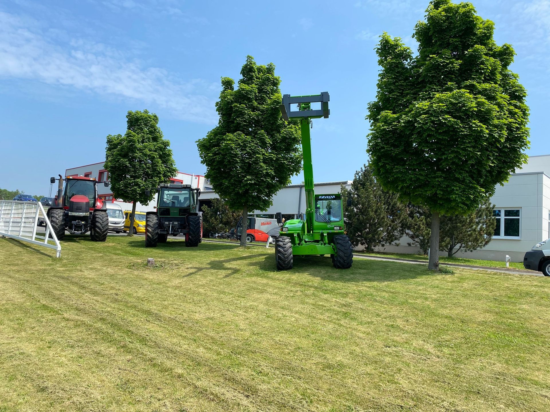 Drei landwirtschaftliche Fahrzeuge (Traktor, Teleskoplader) auf grünem Gras mit Bäumen und einem weißen Gebäude im Hintergrund.