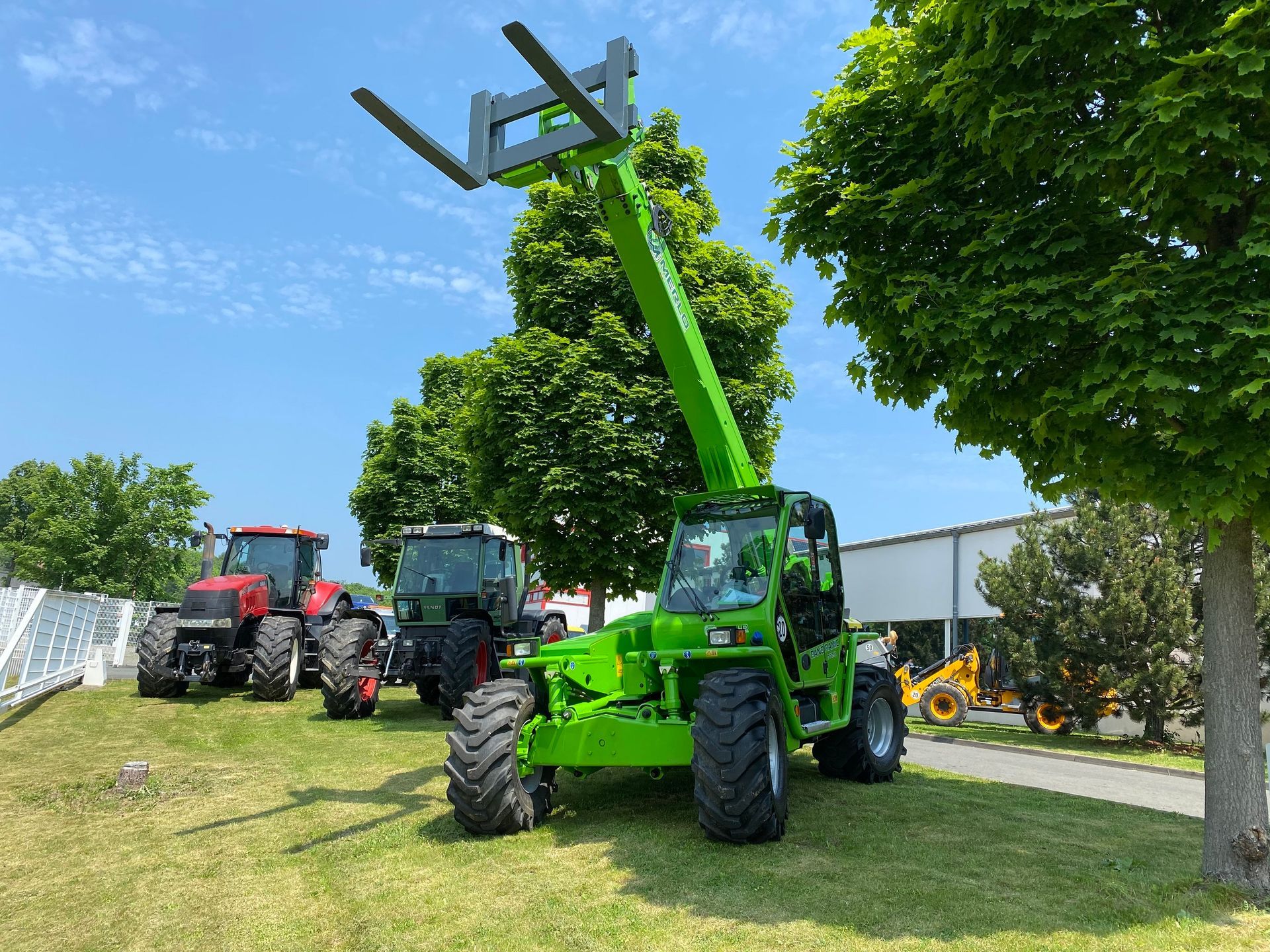 Grüner Teleskoplader mit angehobenen Gabeln; Traktoren und ein blauer Himmel im Hintergrund.