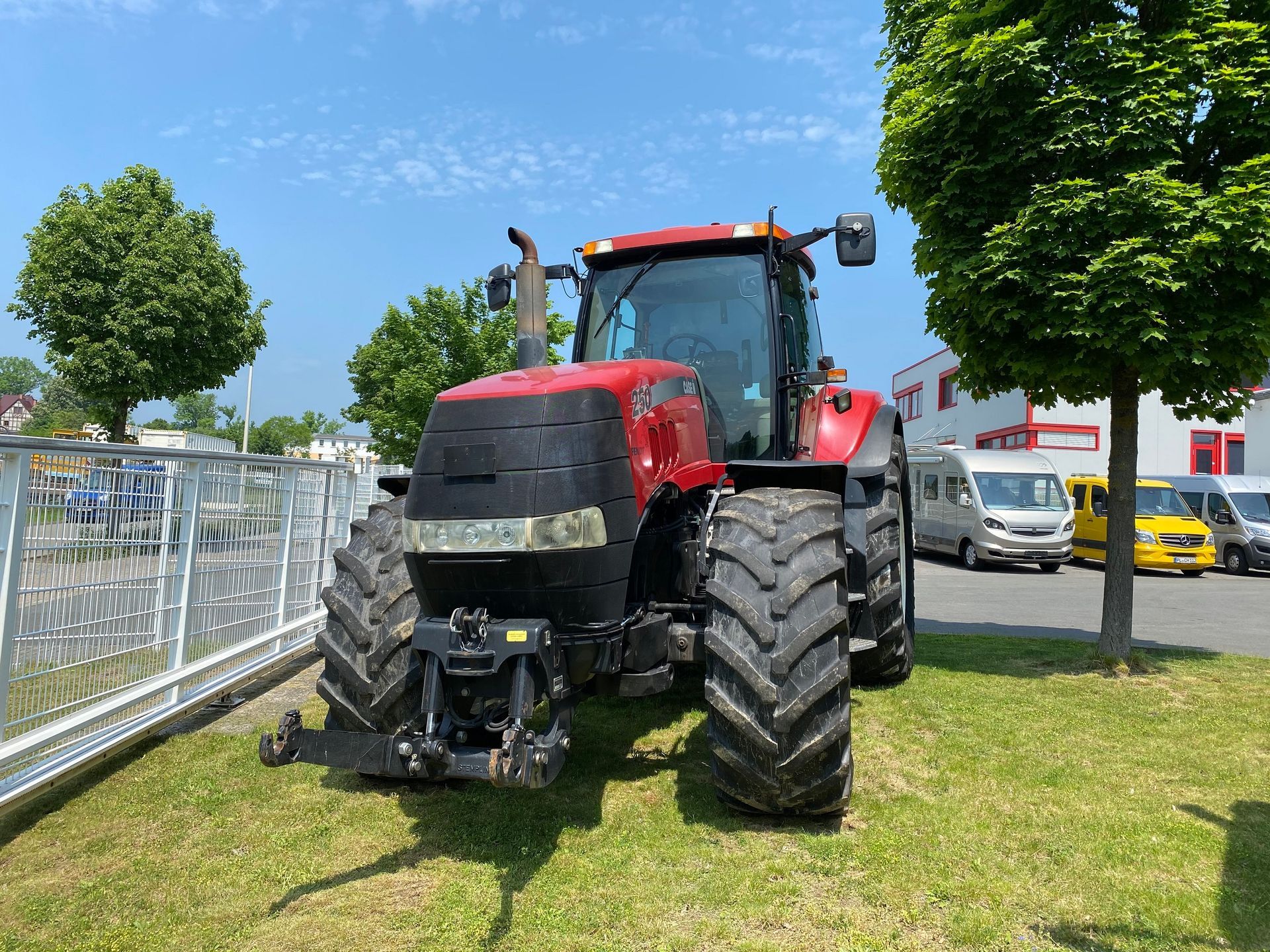 Roter Case IH-Traktor, der an einem sonnigen Tag auf dem Gras vor einem Gebäude und einem Zaun geparkt ist.