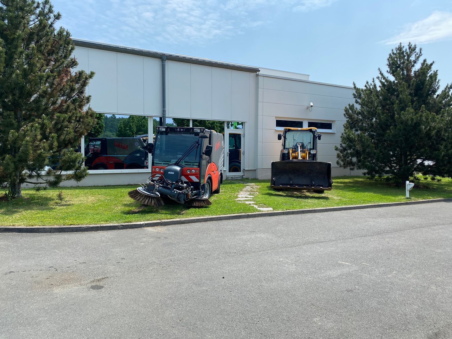 Ein kleines orangefarbenes Fahrzeug und ein gelber Bagger vor einem weißen Gebäude mit grünem Rasen.