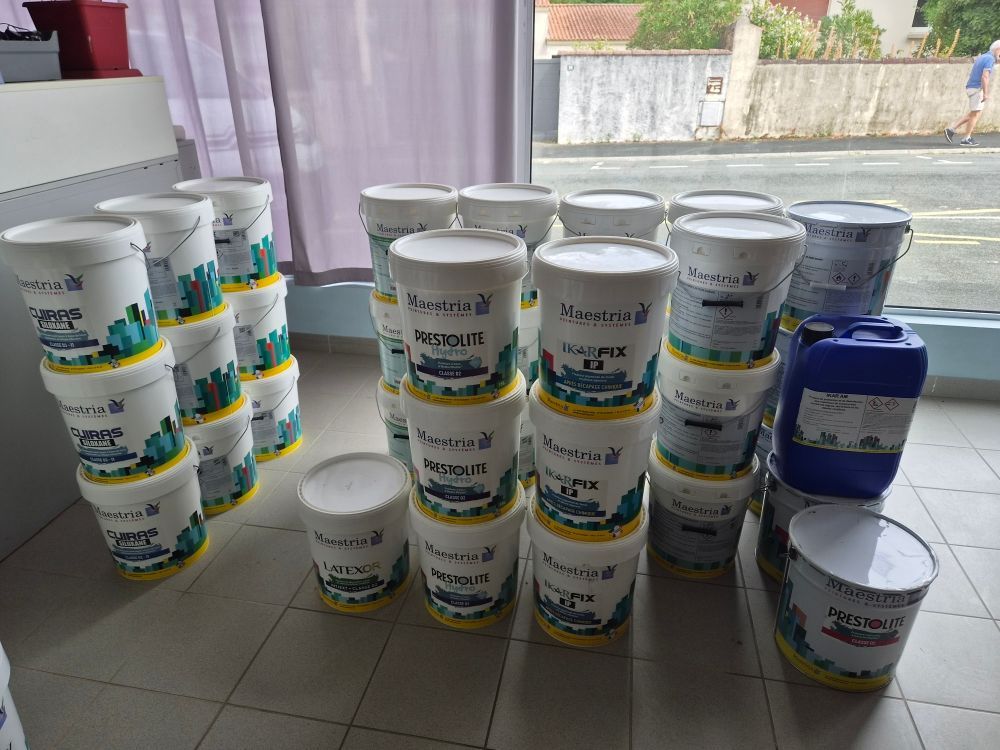 Des piles de seaux de peinture blanche et quelques pots de peinture bleue sont disposés sur un sol carrelé près d'une fenêtre.