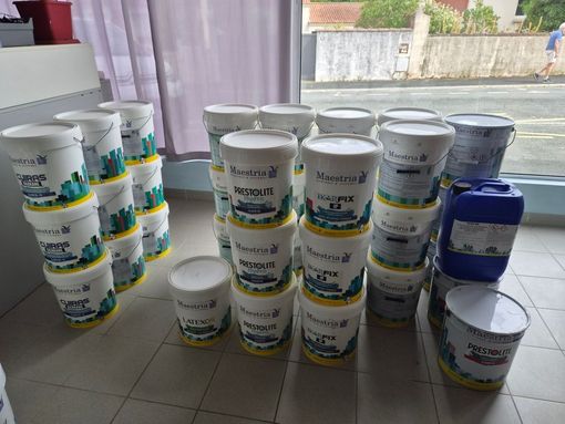 Des piles de seaux de peinture blanche et quelques pots de peinture bleue sont disposés sur un sol carrelé près d'une fenêtre.