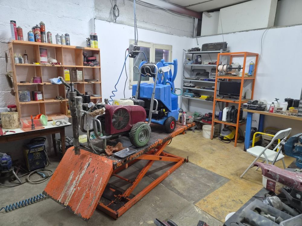 Atelier encombré avec des étagères, des outils et une plateforme orange au premier plan.
