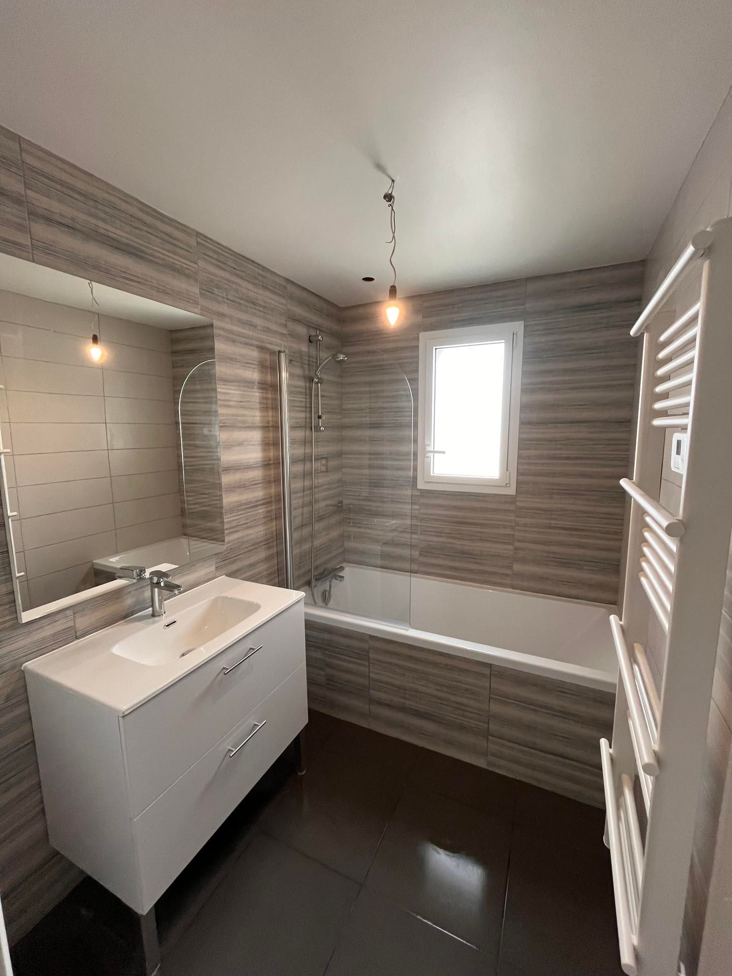 Salle de bains avec meuble lavabo blanc, baignoire, fenêtre et murs carrelés gris.