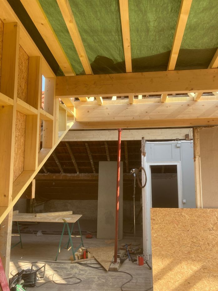 Intérieur à ossature bois en cours de construction, avec escaliers, poutres apparentes et outils et matériaux éparpillés.