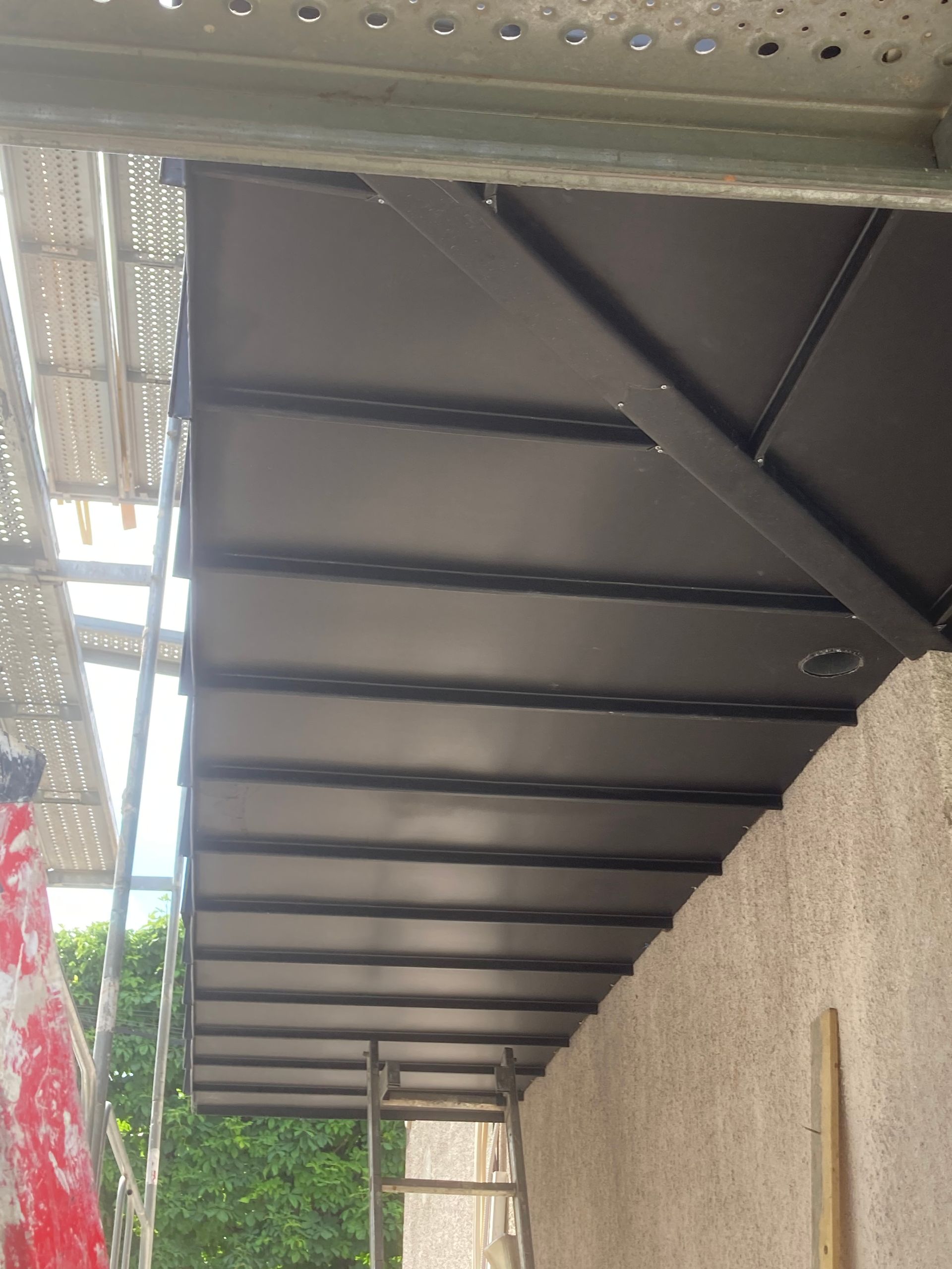 Un auvent noir recouvre une étroite allée extérieure, à côté d'un mur clair et d'un échafaudage.