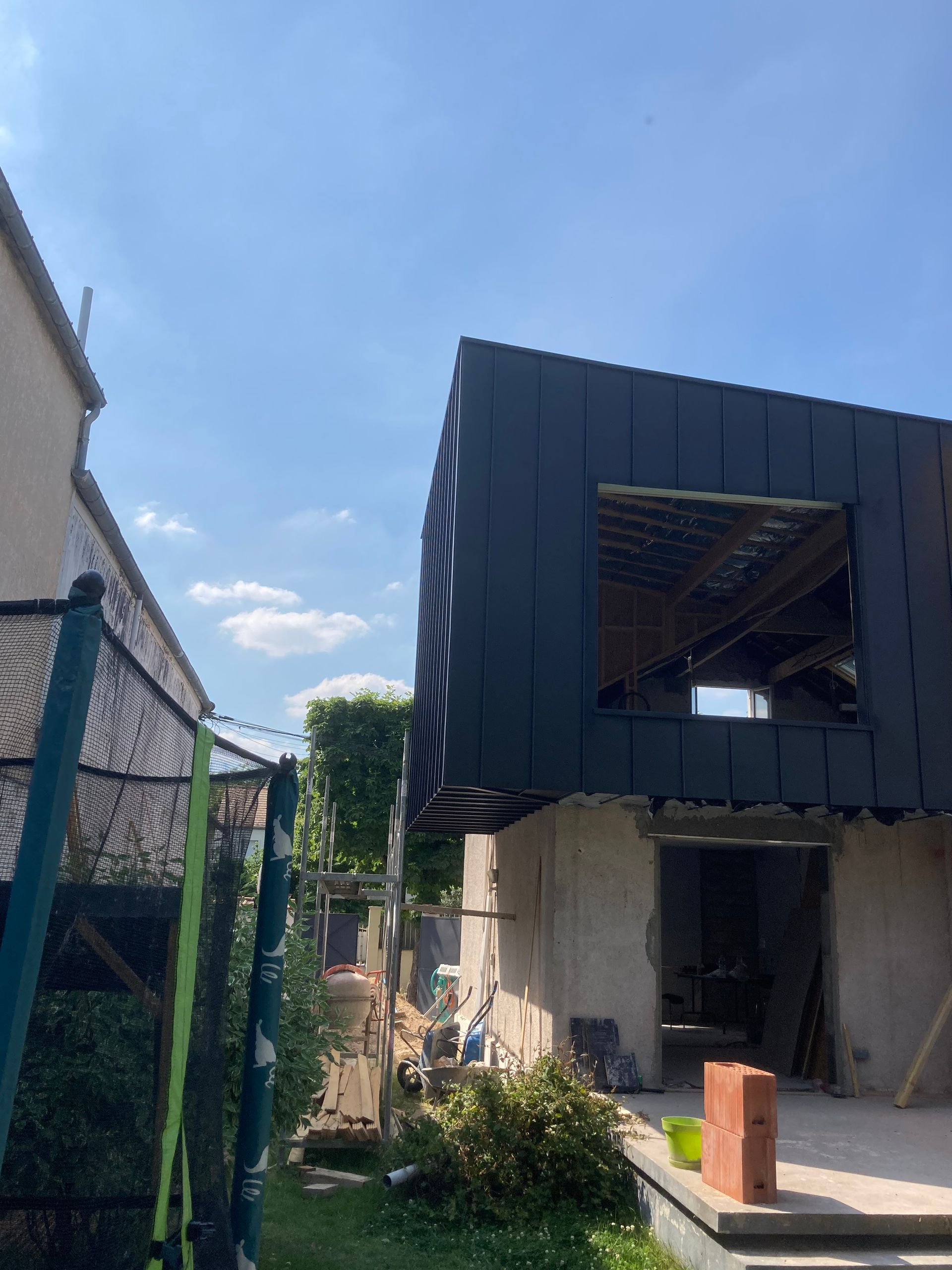 Maison moderne en construction avec façade supérieure sombre, balcon ouvert et petit jardin par une journée ensoleillée