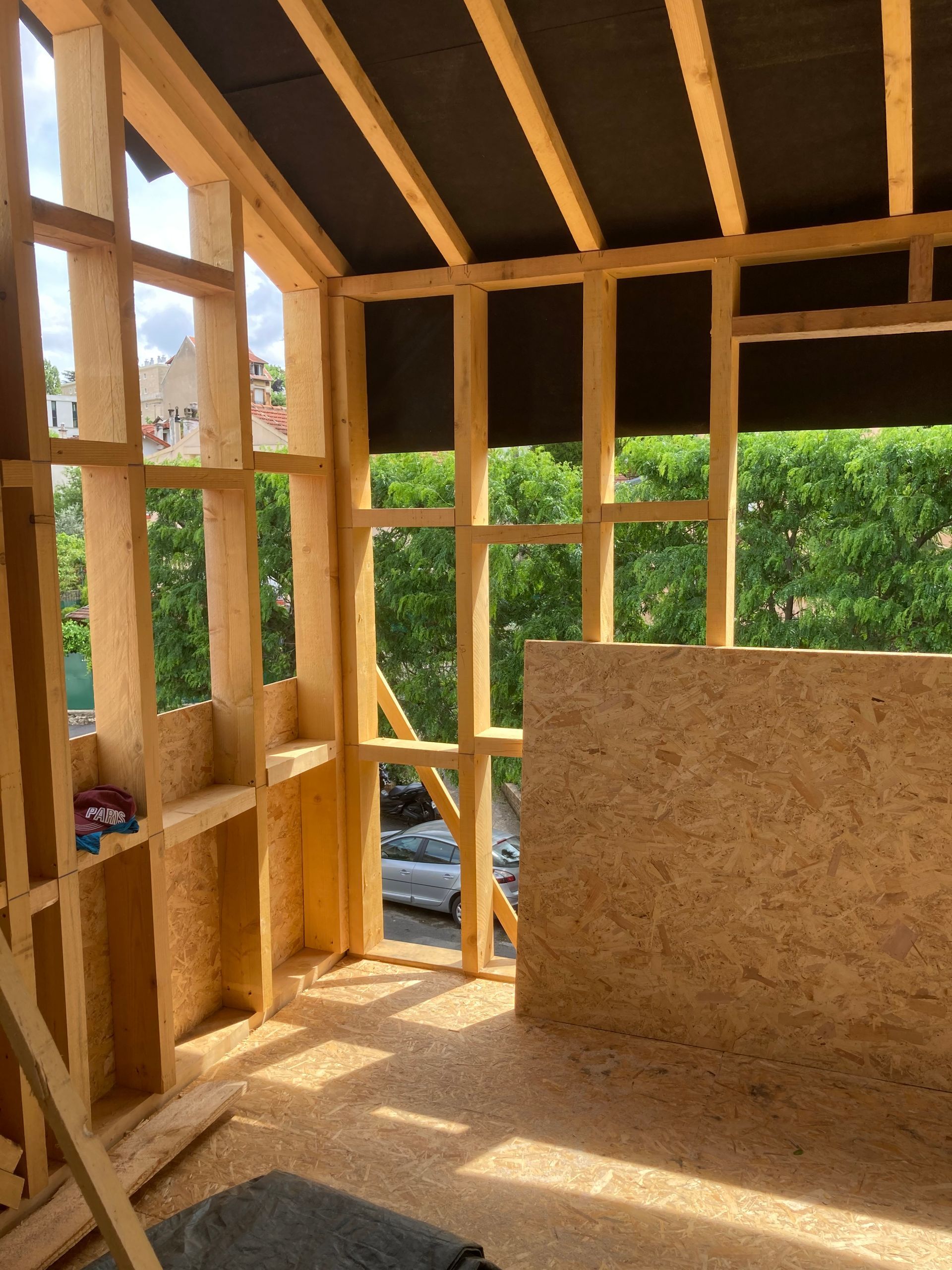 Intérieur d'une pièce à ossature bois en construction, avec des murs en contreplaqué et de grandes fenêtres donnant sur les arbres