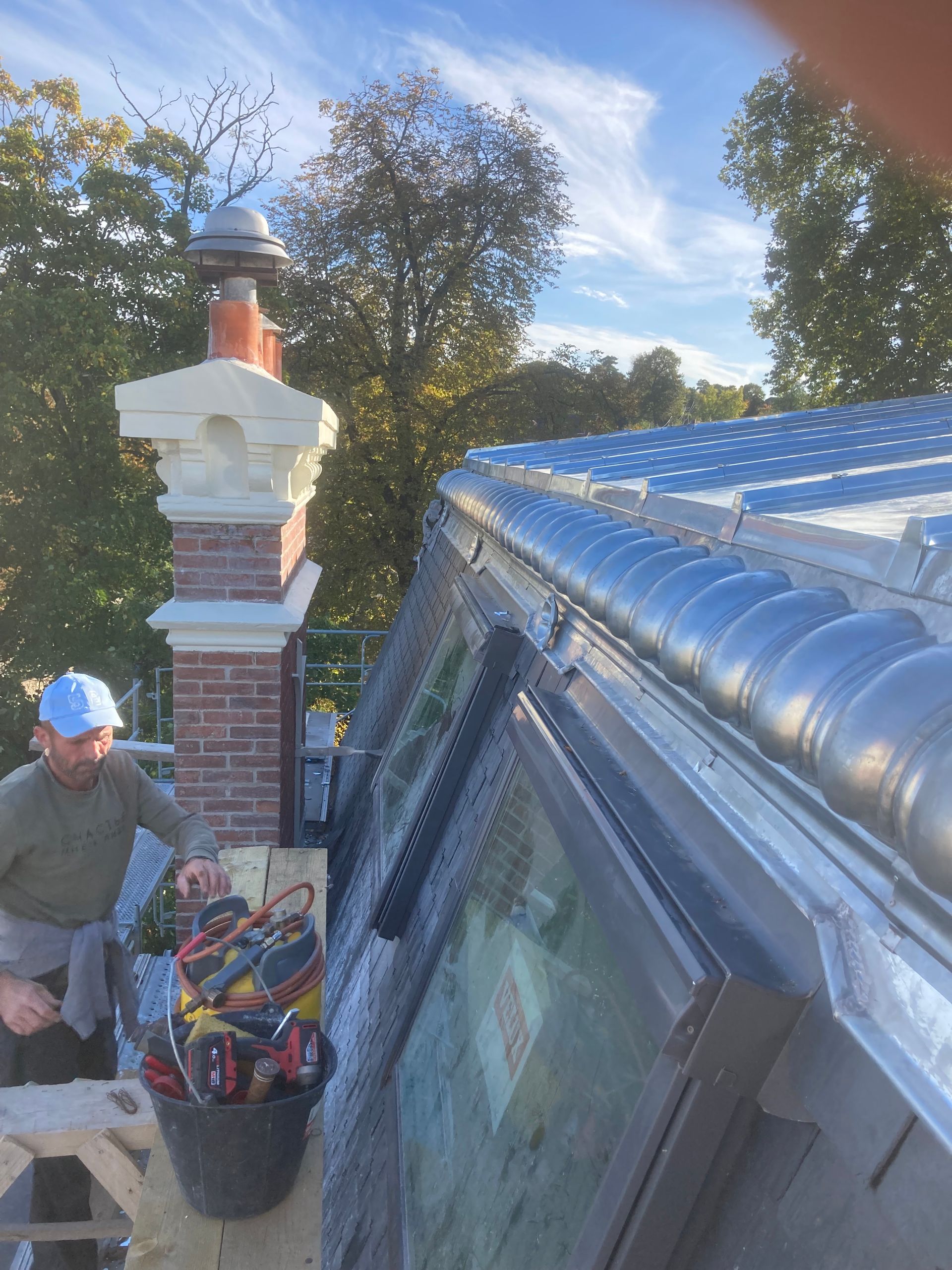 Un ouvrier, sur un toit à côté d'une cheminée en briques, inspecte une toiture métallique réfléchissante sous un ciel bleu.