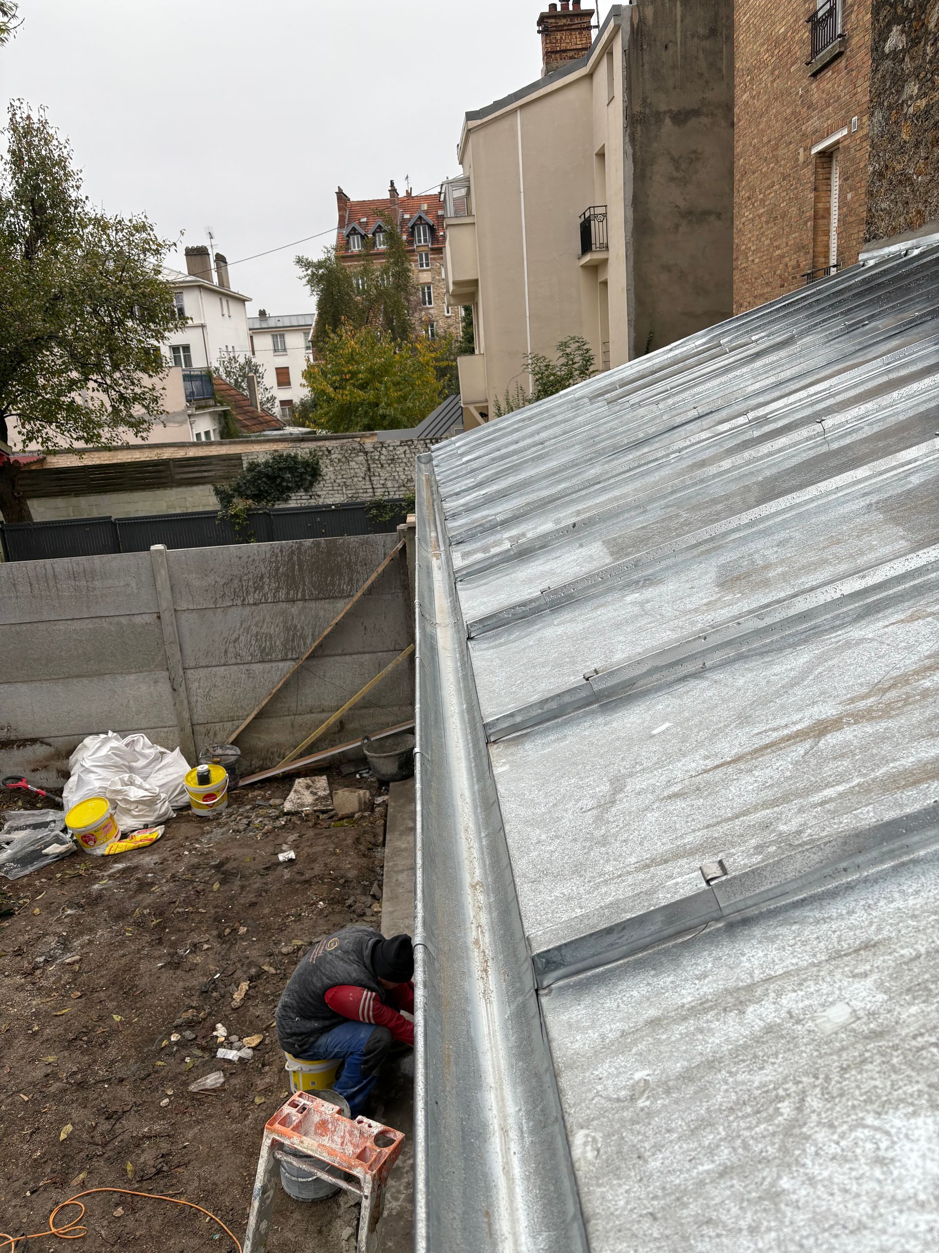 Vue depuis le toit d'une maison en tôle ondulée, donnant sur des jardins et des bâtiments voisins en brique et en stuc.