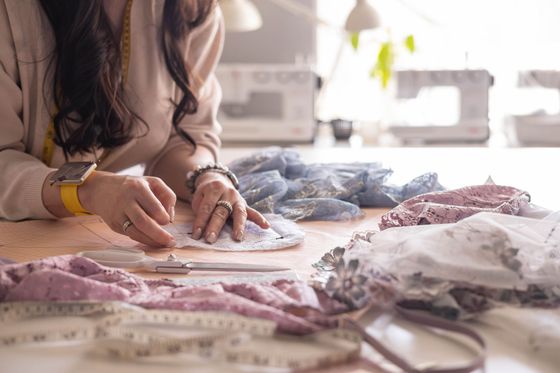 Mujer cose tela de encaje en una mesa con máquina de coser, rodeada de tela y herramientas de medición.