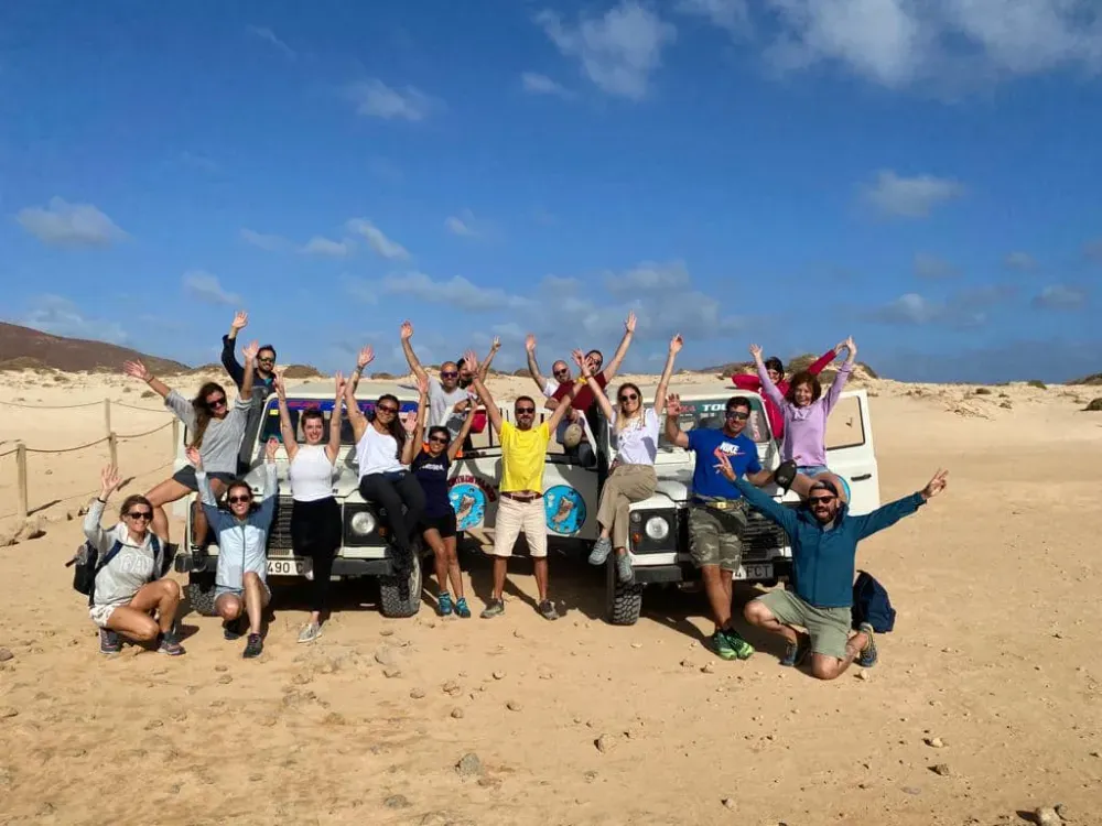 Un numeroso grupo de personas posa con los brazos en alto frente a dos vehículos todoterreno blancos en un paisaje desértico arenoso.