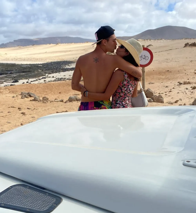 Una pareja se besa apoyada en el capó de un vehículo blanco en un paisaje soleado que parece desértico.