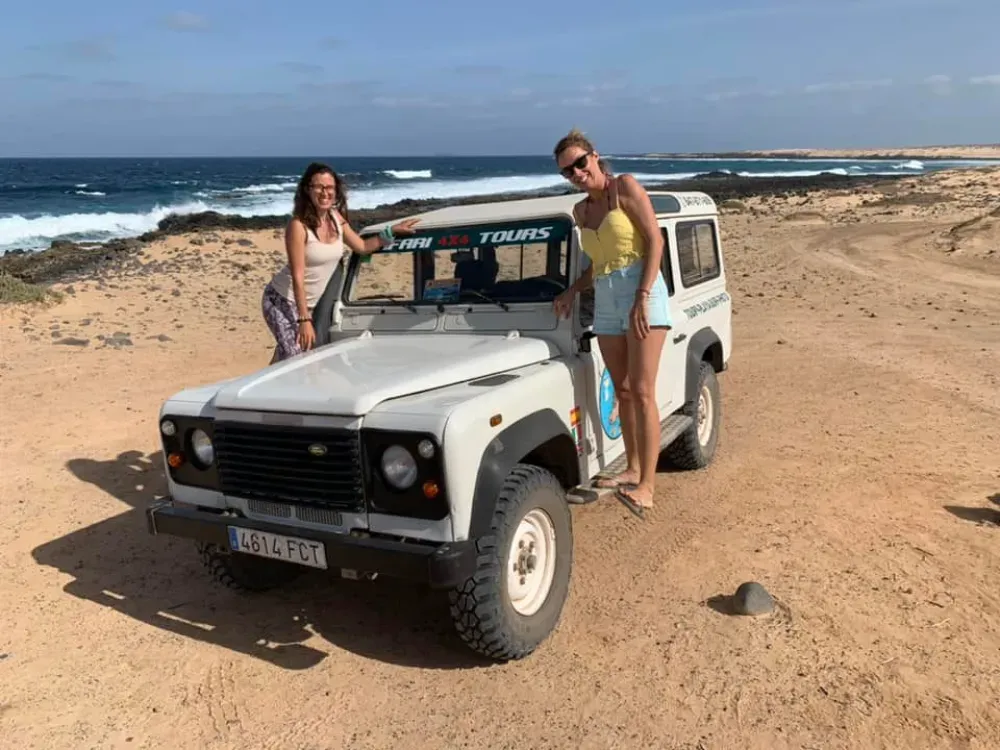 Dos personas sonríen mientras posan junto a un Land Rover blanco estacionado en una costa desértica con vistas al mar.