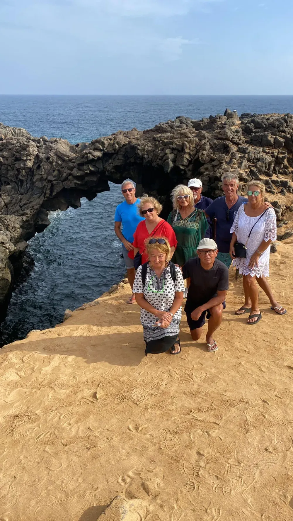 Un grupo de personas de pie sobre un acantilado rocoso de color dorado, junto a un arco natural de piedra volcánica con vistas al océano.