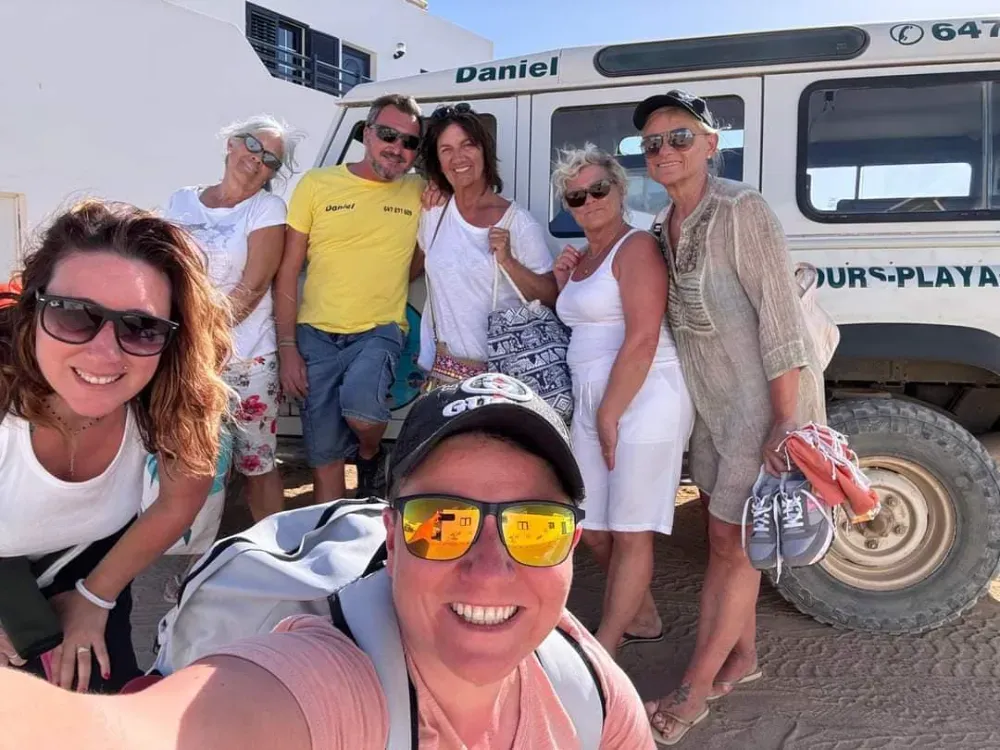 Un grupo de siete personas sonriendo en una selfie al aire libre frente a un vehículo todoterreno blanco.