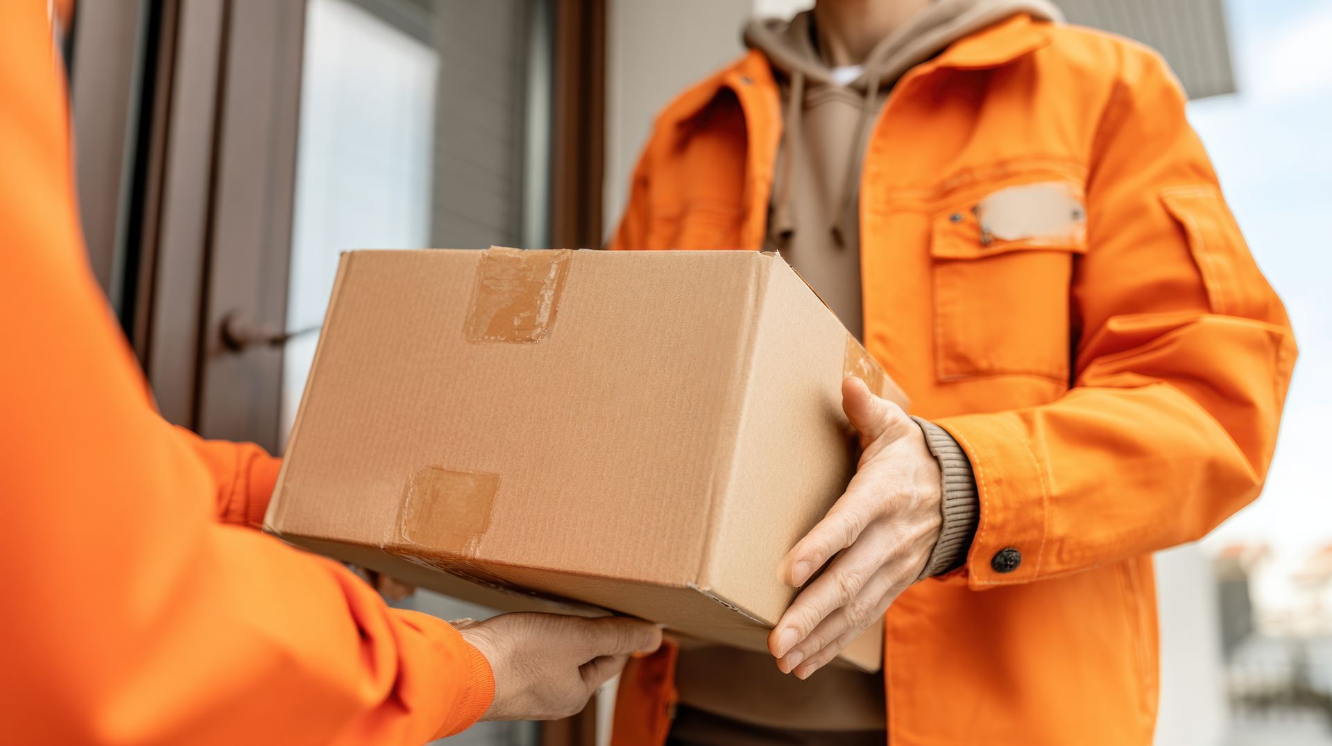 Un livreur en veste orange remet un carton sur le pas d’une porte.