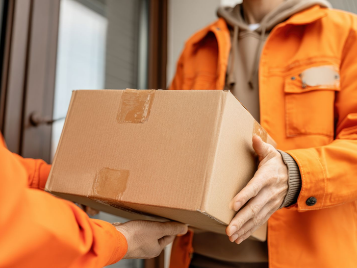 Personne en veste orange remettant un carton à une autre personne sur le pas d’une porte.