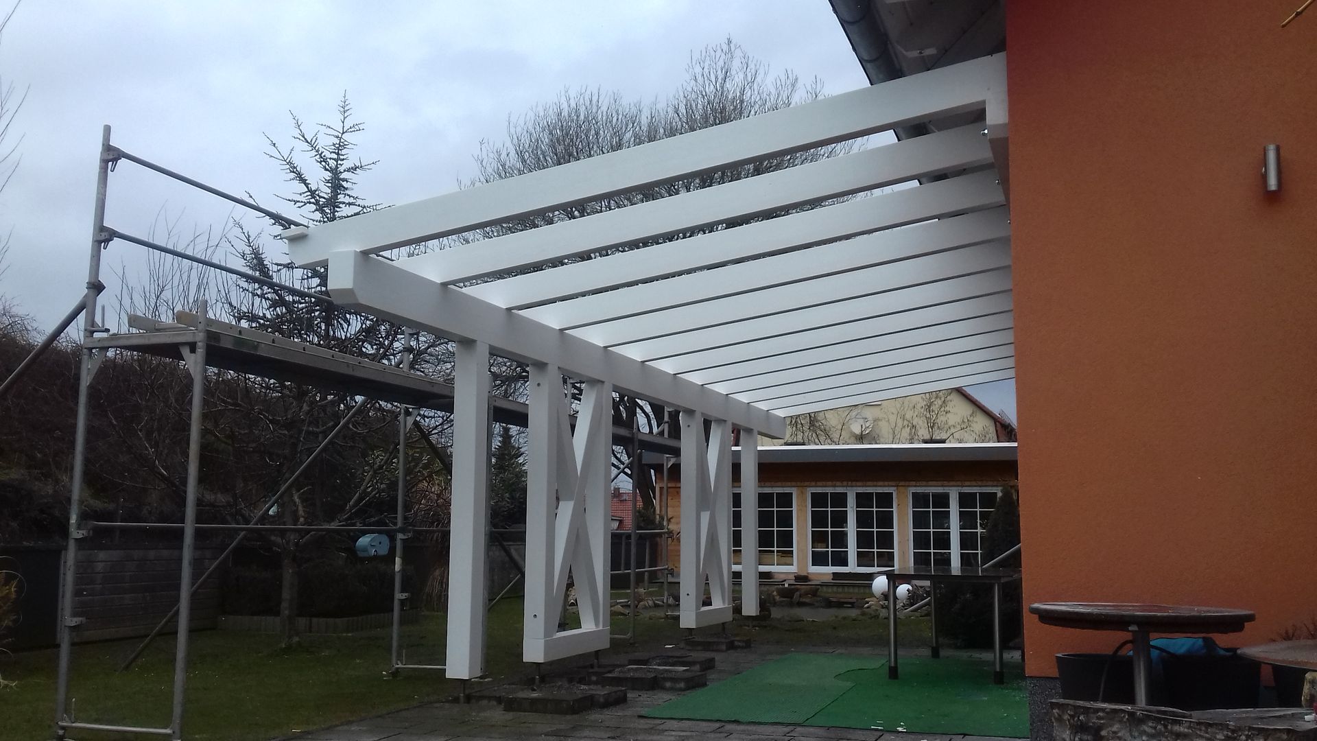 Weiße Holz-Pergola im Garten vor einem Wohnhaus mit überdachter Terrasse und Querbalken.