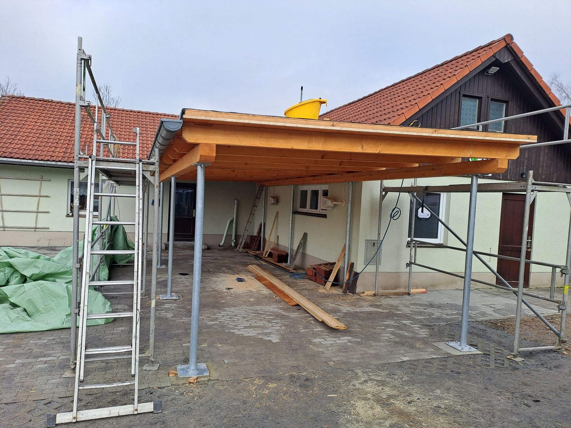 Überdachte Terrasse im Bau mit Holzdachkonstruktion und Gerüst vor einem Wohnhaus.
