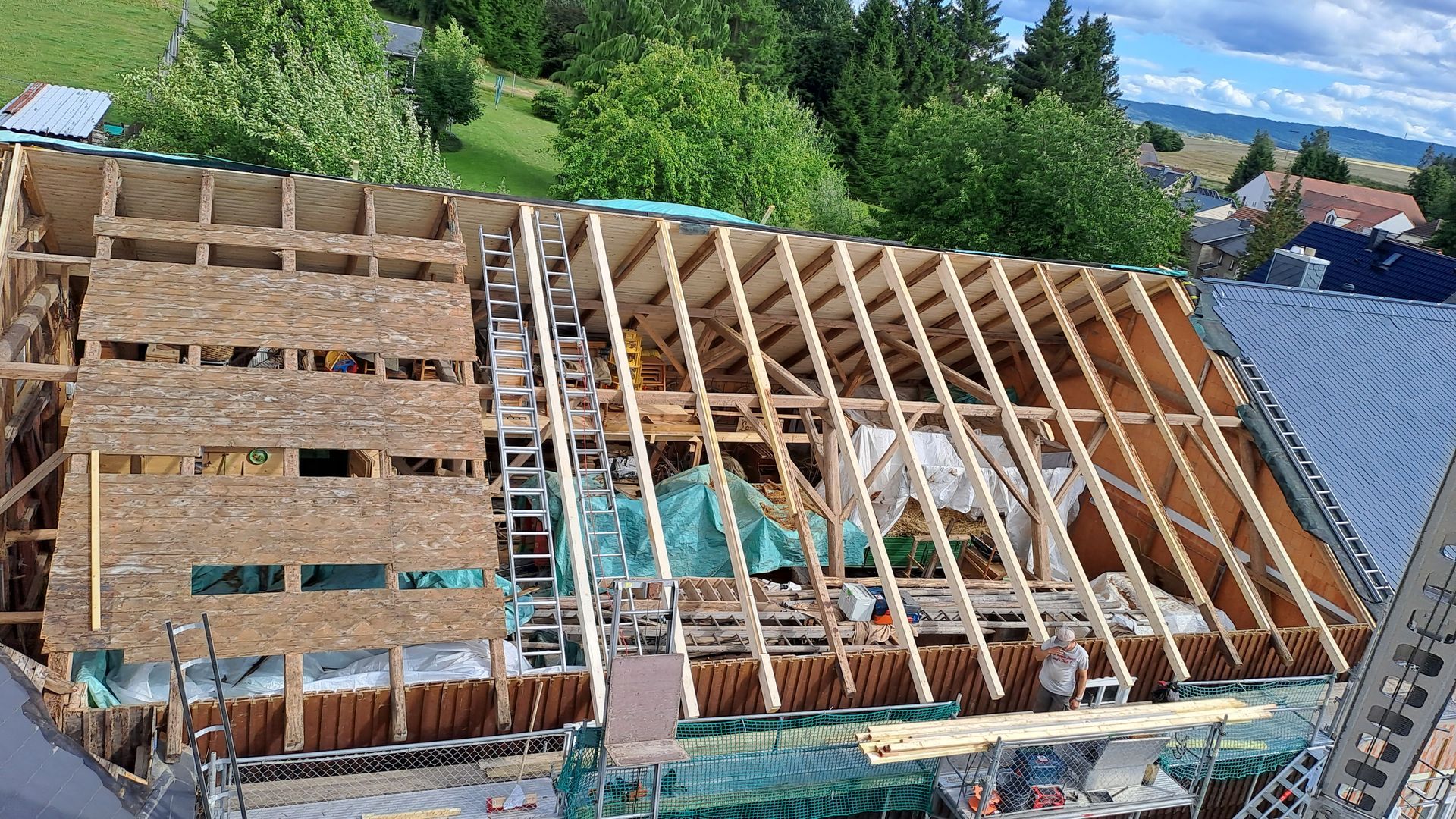 Baustelle mit neu errichtetem Dachstuhl aus Holz, Gerüst und Leiter an einem Hanggrundstück.