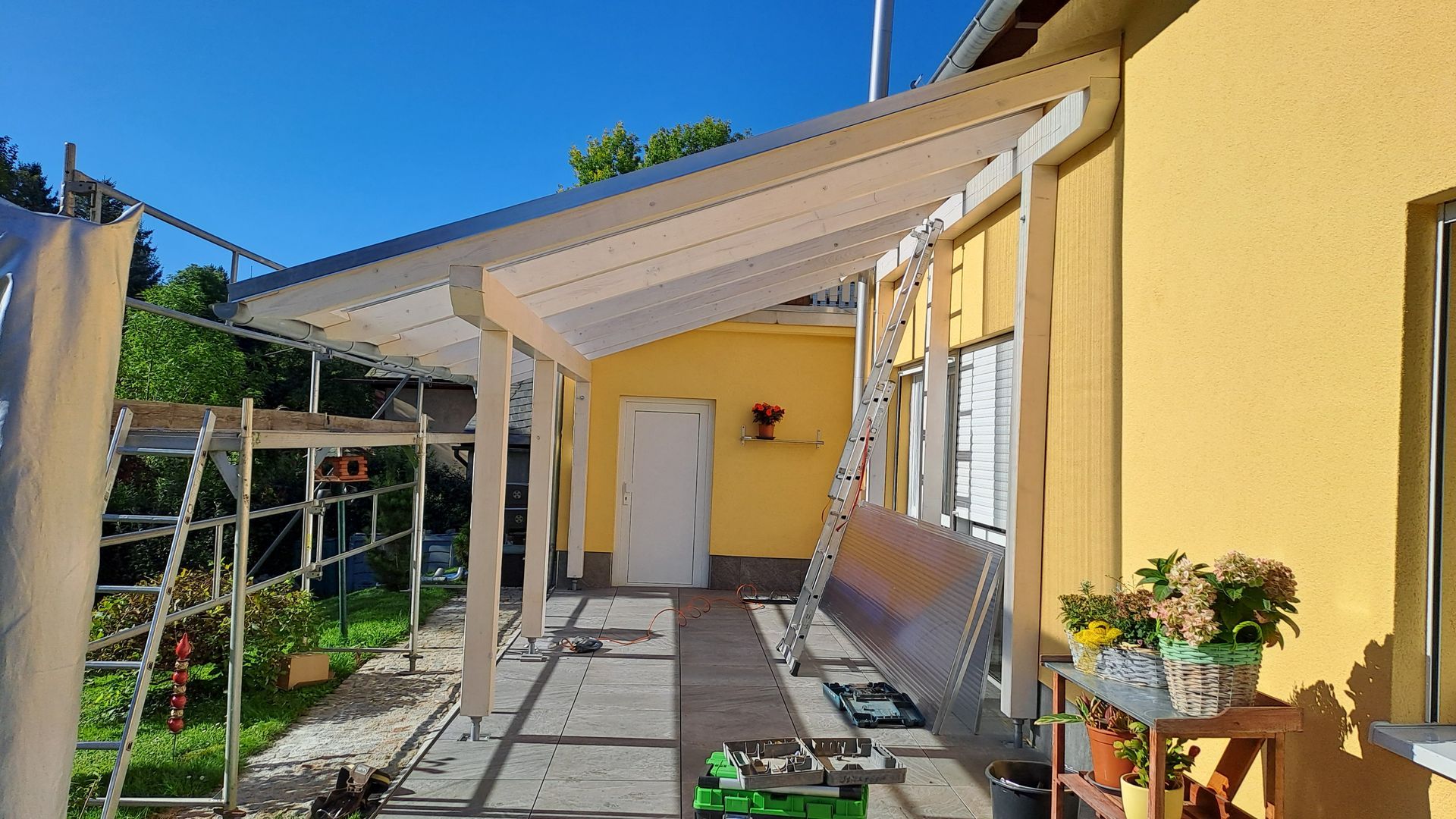 Überdachte Terrasse im Bau mit weißer Holzkonstruktion und transparenten Dachplatten an einem gelben Haus.