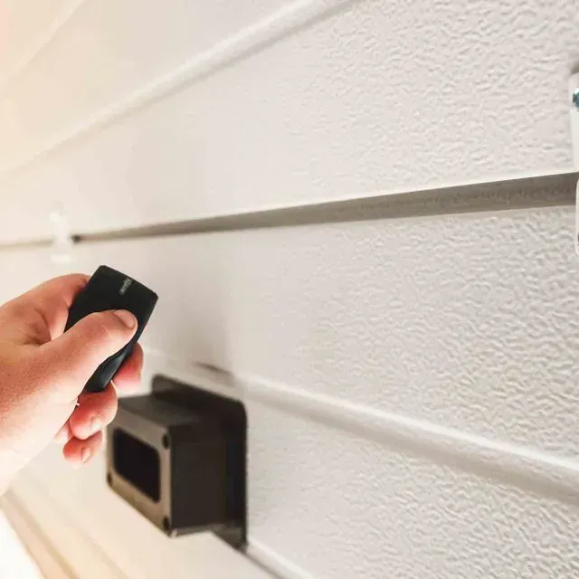 Una persona sostiene un control remoto frente a una pared blanca.