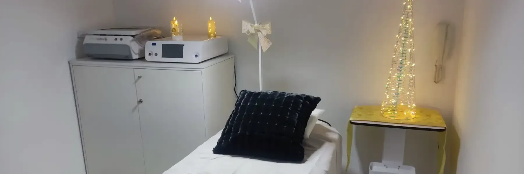 Una sala de tratamiento limpia con una camilla de masaje blanca, un cojín negro, equipo médico en un armario