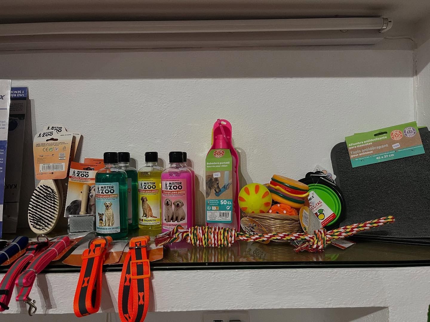 Suministros para mascotas en un estante: cepillos, champús, juguetes y correas en varios colores, contra una pared blanca.