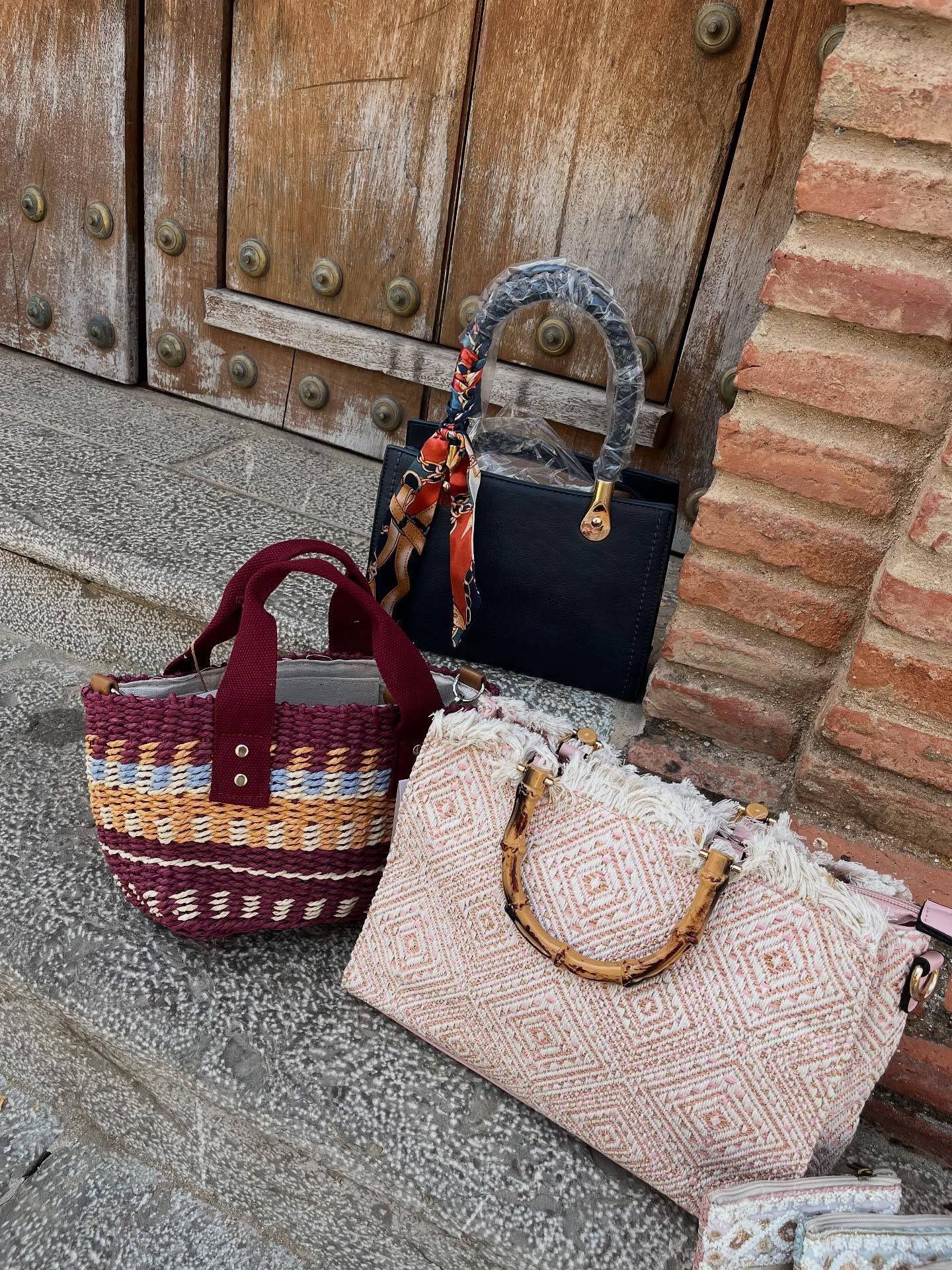 Tres bolsos sobre una superficie de piedra: un tote tejido en color rojo, un satchel estructurado en color negro y un bolso de tela en color beige.