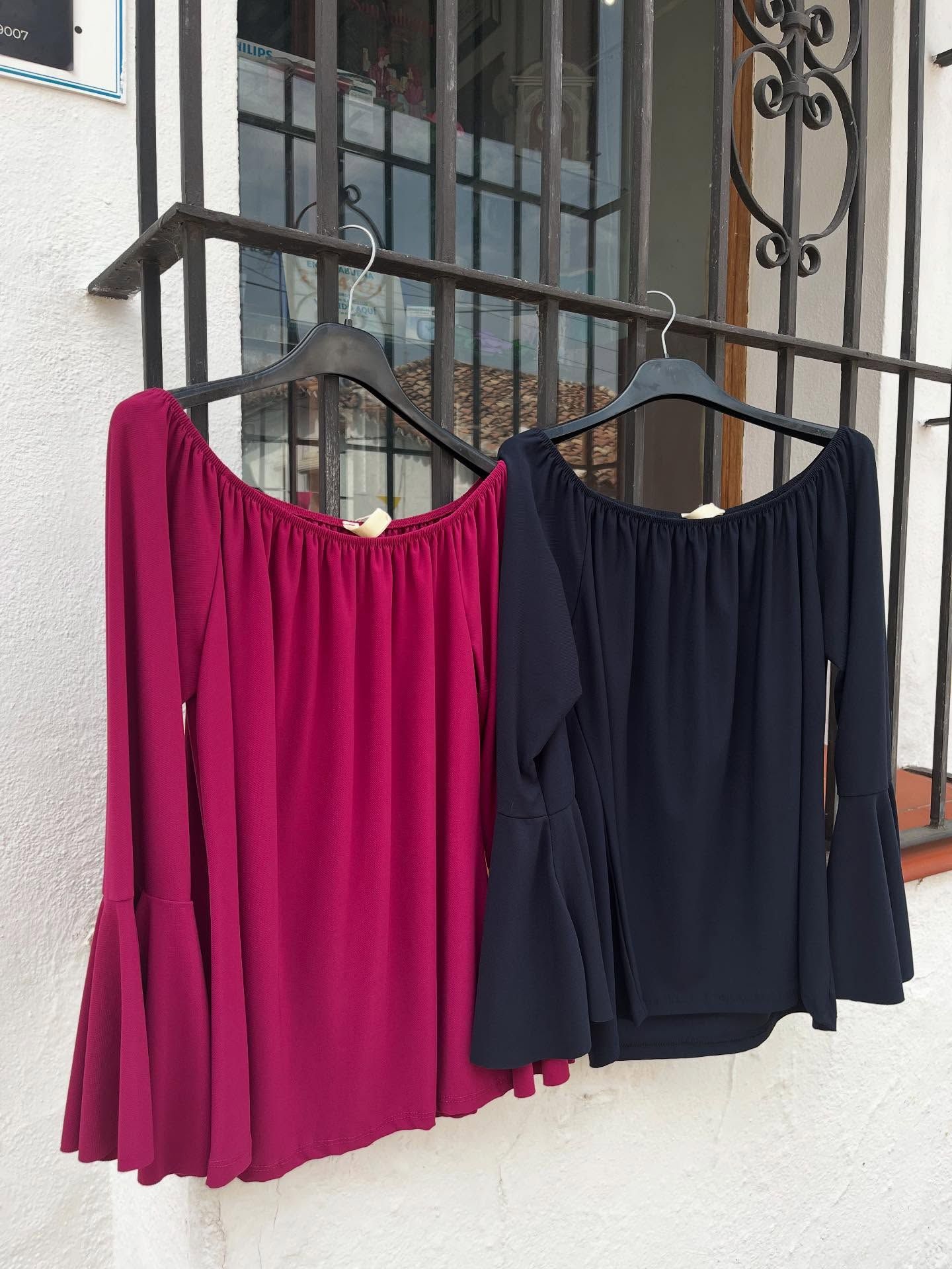 Dos tops con hombros descubiertos, en color magenta y azul marino, con mangas acampanadas, colgados de una barandilla.