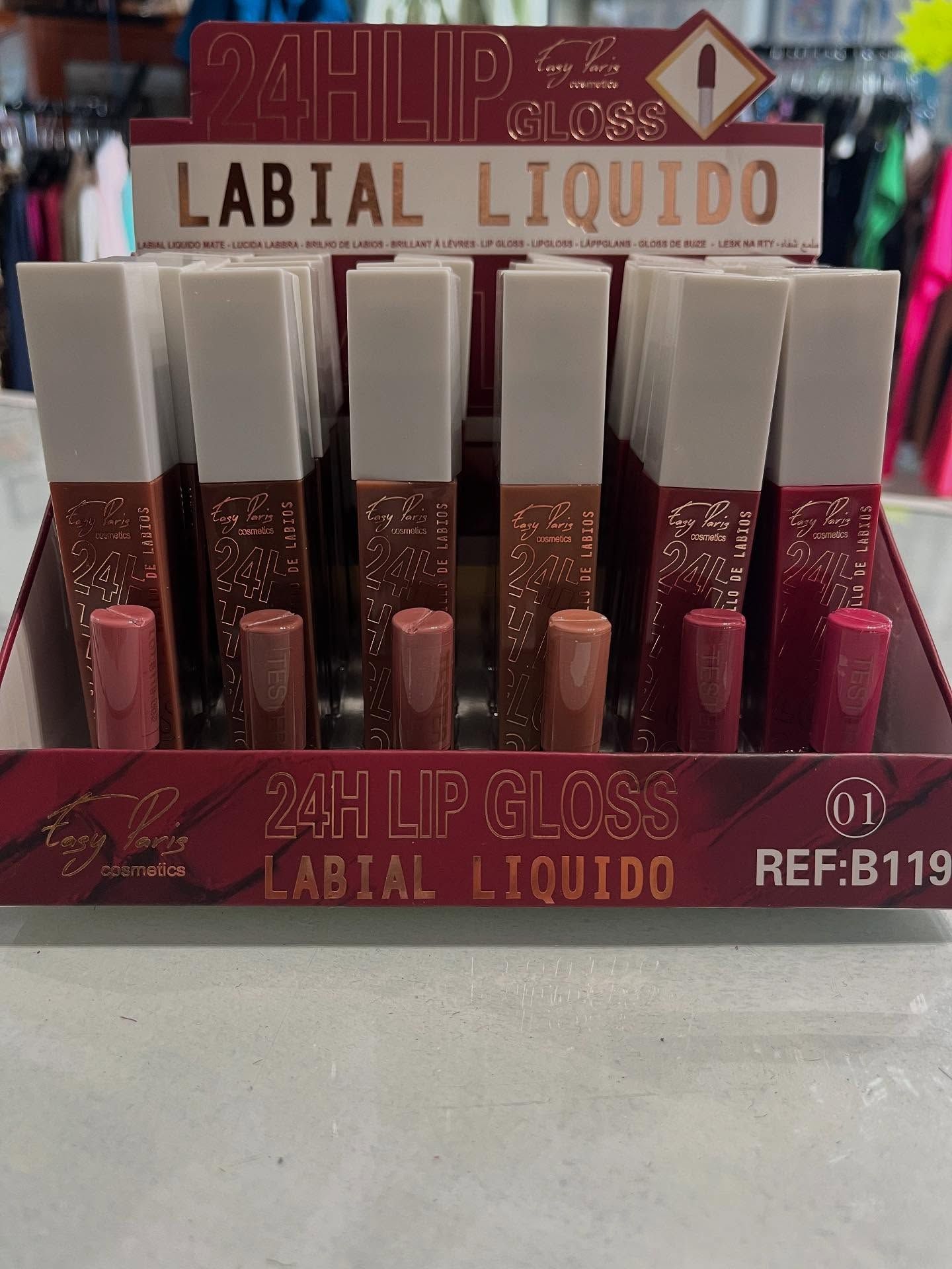 Expositor de brillo labial líquido 24H en varios tonos, packaging blanco y dorado.